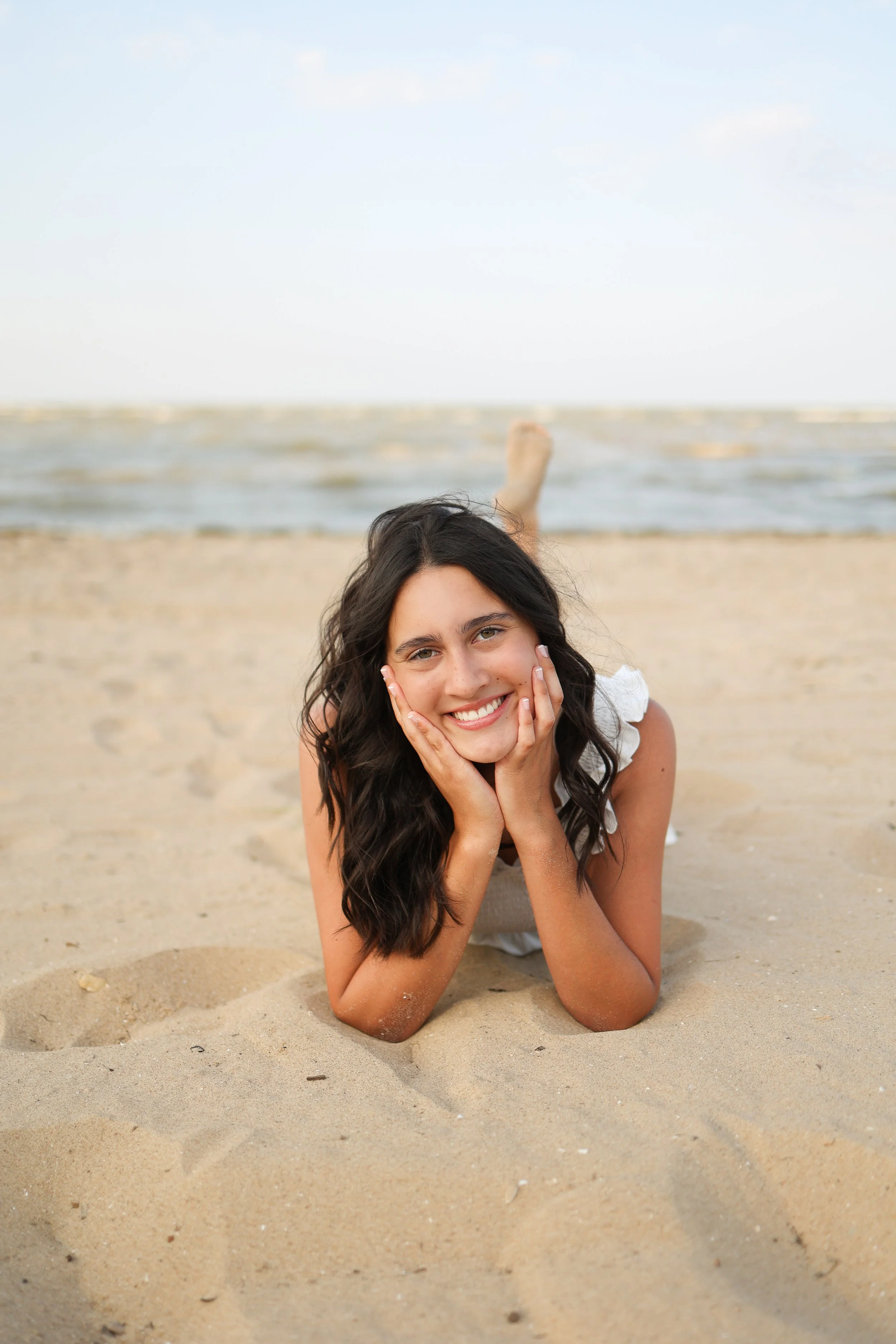 A woman lying on a sandy beach, propped up on her elbows, smiling with her hands on her cheeks. The ocean and sky are visible in the background.