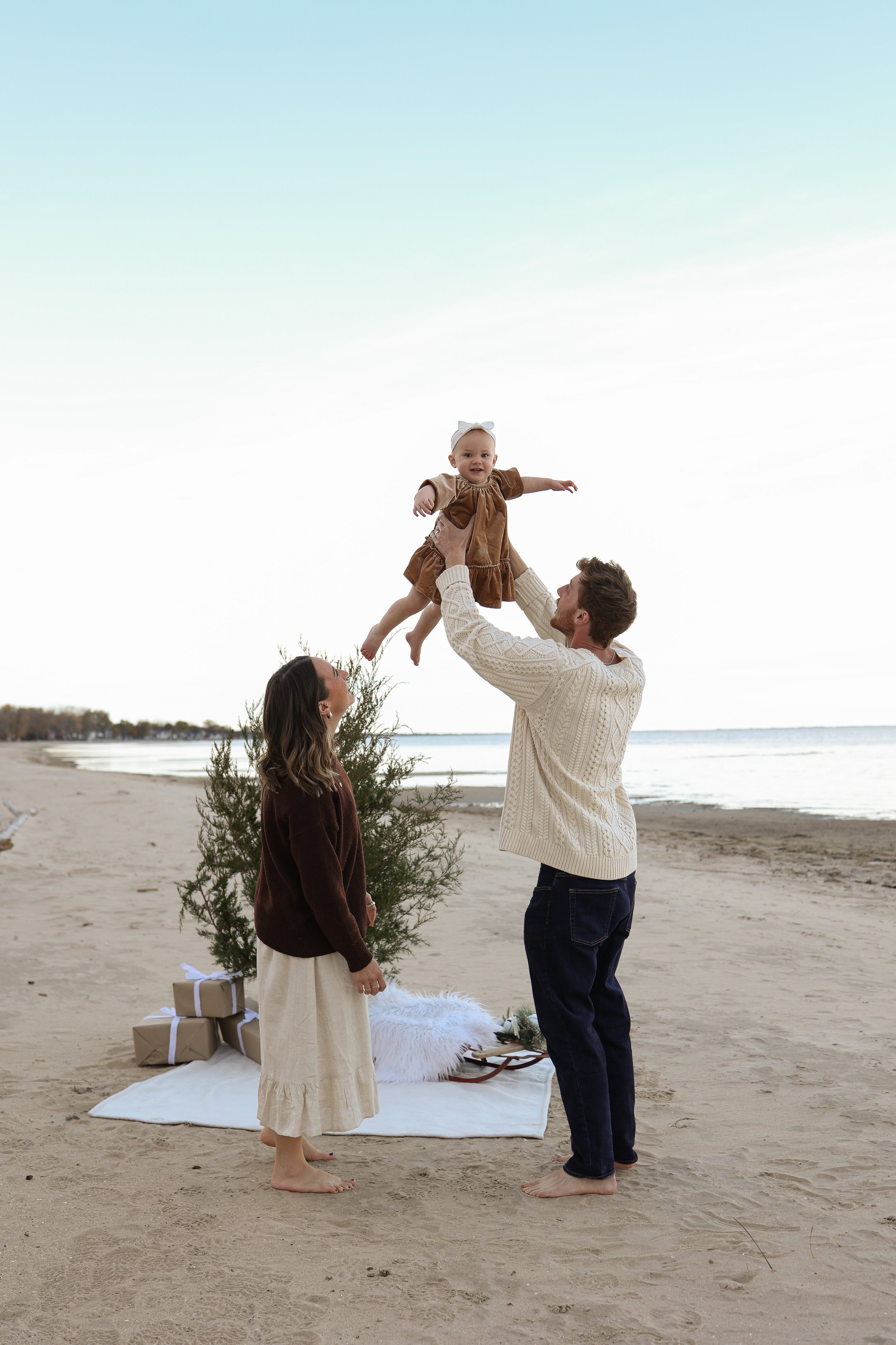 A family celebrating Christmas on the beach, with a man and woman lifting a smiling baby girl, with presents and a small decorated tree on a white blanket.