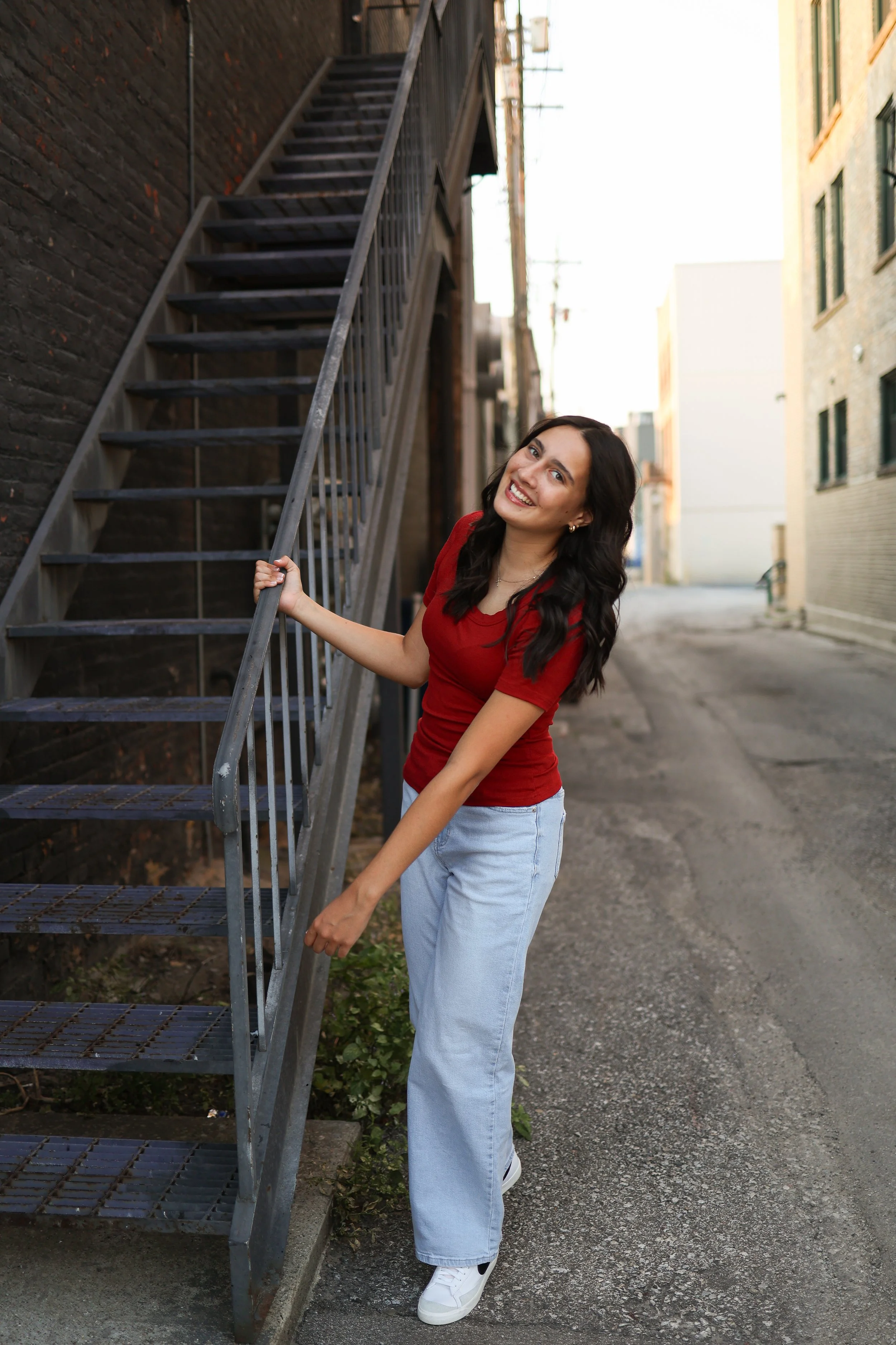 A young woman with long dark hair, wearing a red shirt and light blue jeans, smiling and holding onto a metal staircase railing in an urban alleyway.