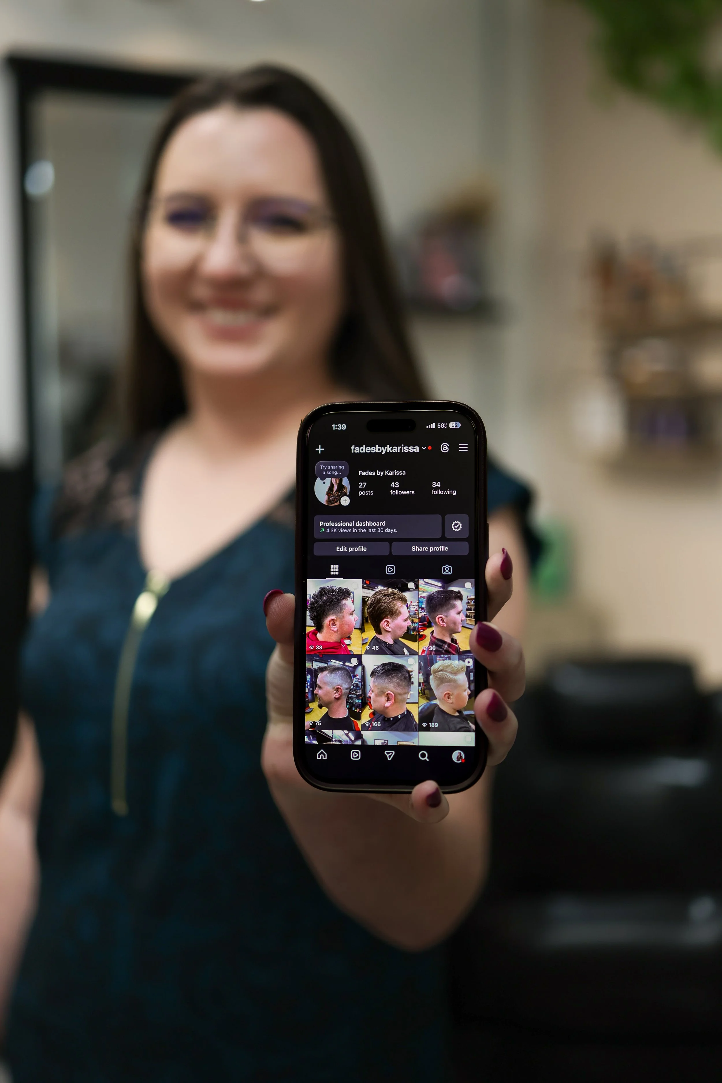 A woman holding a smartphone displaying her Instagram profile with hair styling photos to show branding, in a blurred indoor setting.