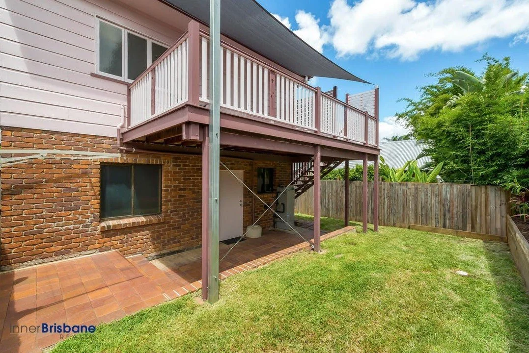 Ground view of the backyard of a house showing a brick wall, a window, a small tiled patio, and an elevated wooden deck with stairs, surrounded by a green lawn and tall trees, under a partly cloudy sky.