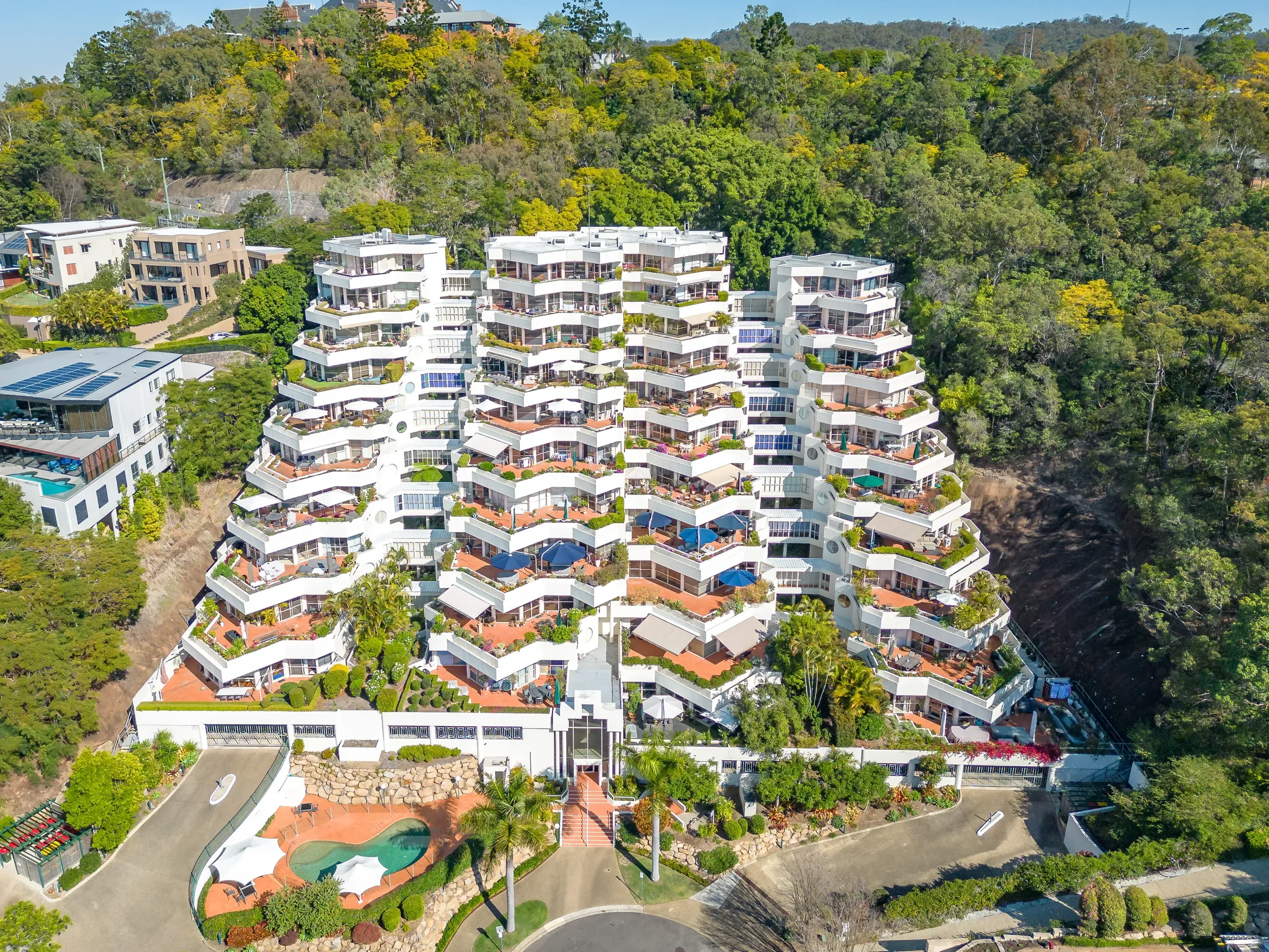 Aerial view of a large multi-story apartment complex on a hillside with lush green trees, outdoor terraces, and a small swimming pool in the foreground.