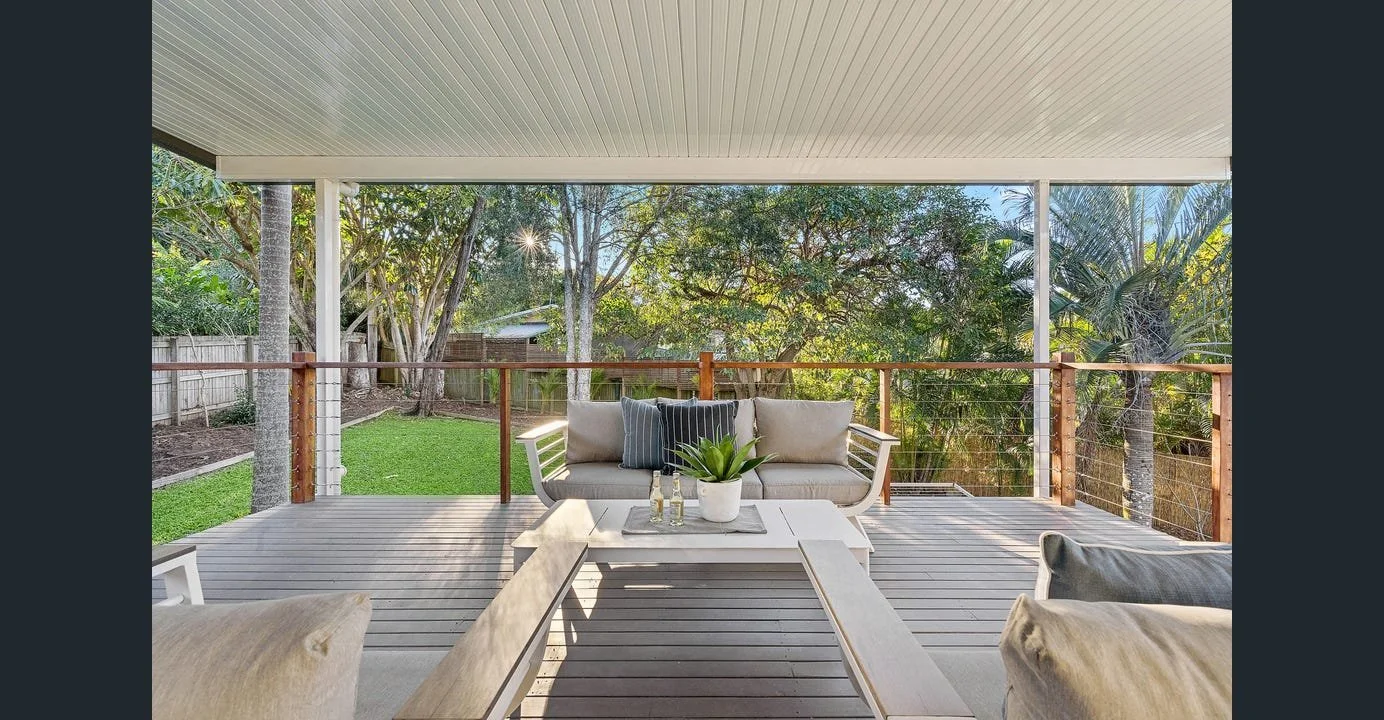 Backyard wooden deck with outdoor furniture, including a couch and chairs with cushions, a coffee table with a potted plant, and a railing with trees in the background.