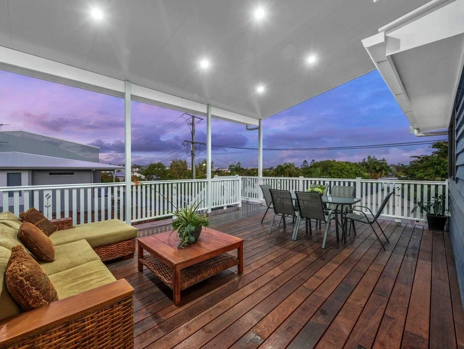 Covered outdoor deck with wicker sofa and pillows, wooden coffee table with plant, metal dining table with four chairs, potted plants, and a view of trees and power lines under a purple sky.