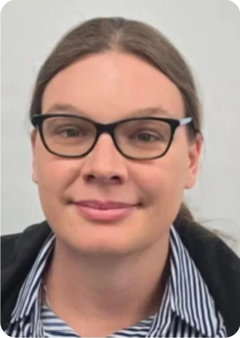 A close-up of a smiling person with long brown hair, wearing black-rimmed glasses, a striped collared shirt, and a dark jacket, against a plain background.