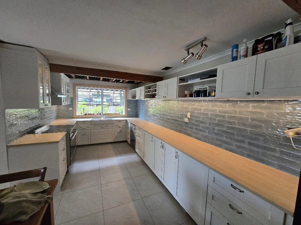 Empty kitchen with white cabinets, gray tile backsplash, and wooden countertops, located near a window with a view outside.