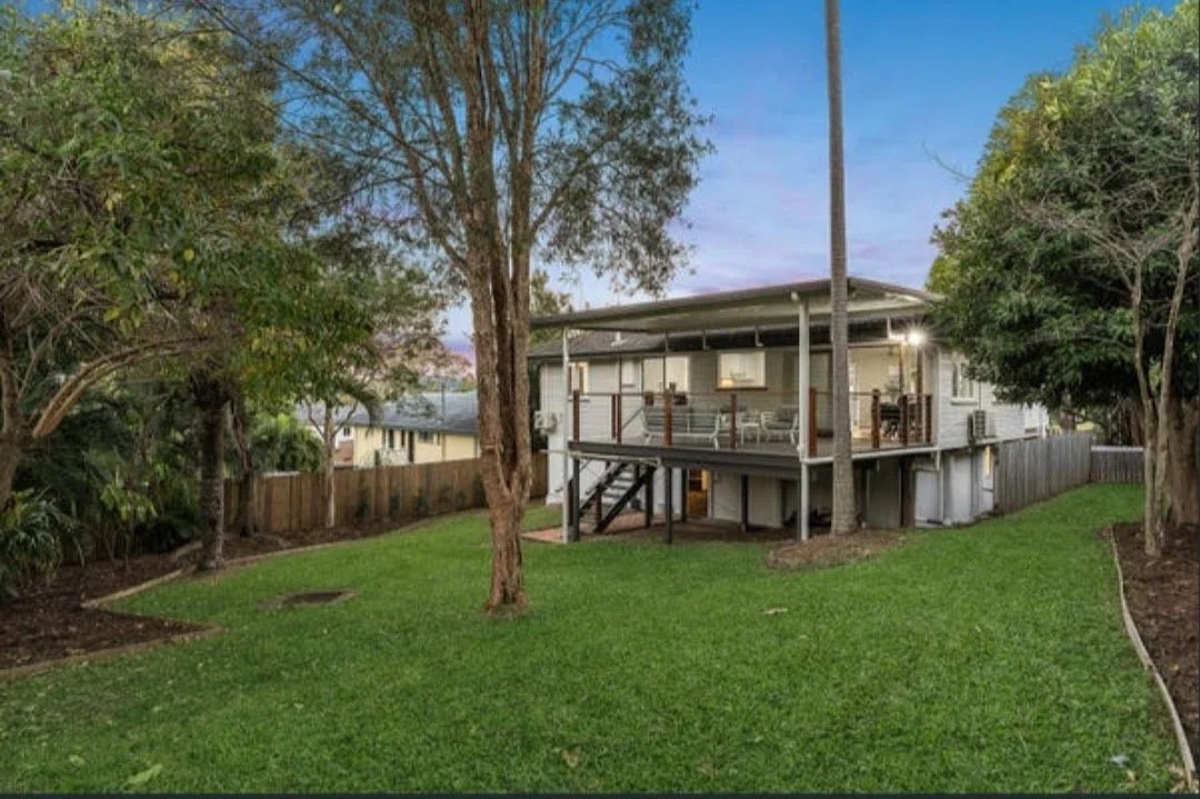 A house with a large backyard, trees, and a wooden deck with stairs, in a suburban neighborhood during dusk.