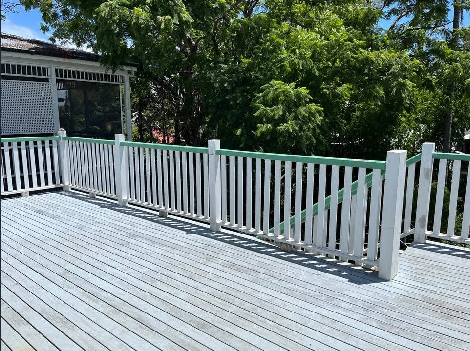 Sunlit wooden deck with white railings and a staircase leading down, surrounded by lush green trees.