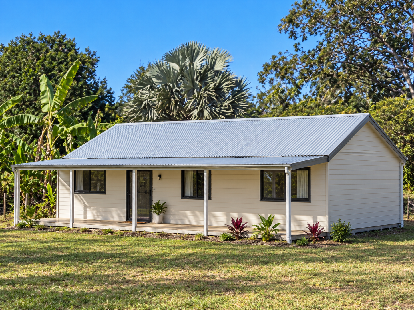A small, beige house with a metal roof, three windows, a door, and a small porch with plants, surrounded by grass and trees under a blue sky.