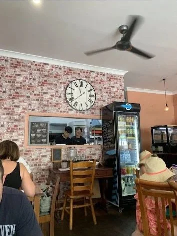 People sitting at tables in a cafe with a brick wall, a large clock, and a vending machine in the background.