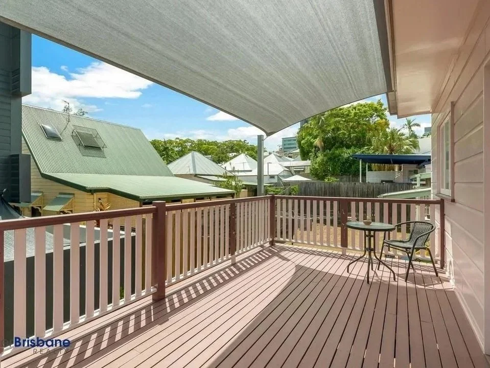 View of a residential balcony with pink railing and wooden flooring, shaded by a sunshade, overlooking neighboring houses and trees.