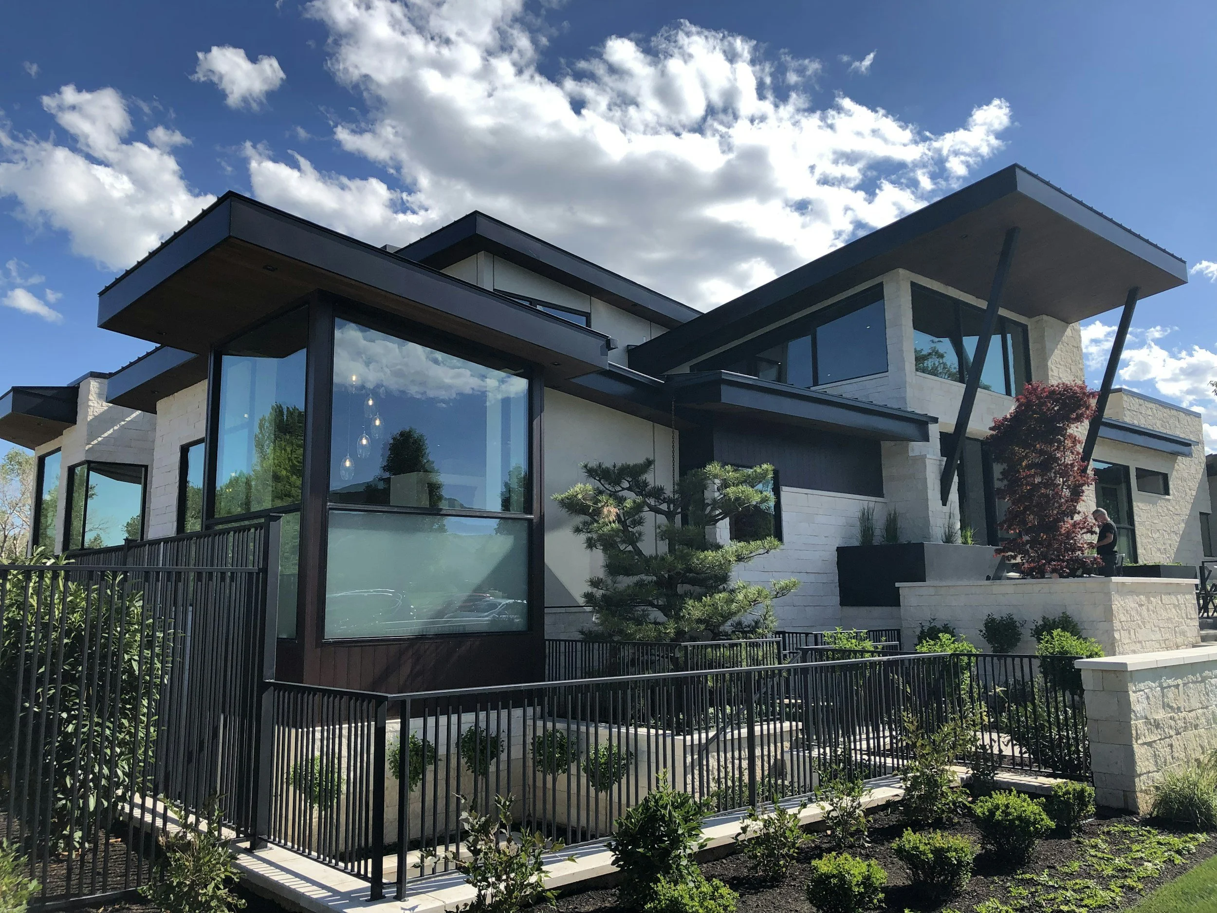 Modern multi-story house with large windows, black accents, and landscaped garden under a blue sky with clouds.