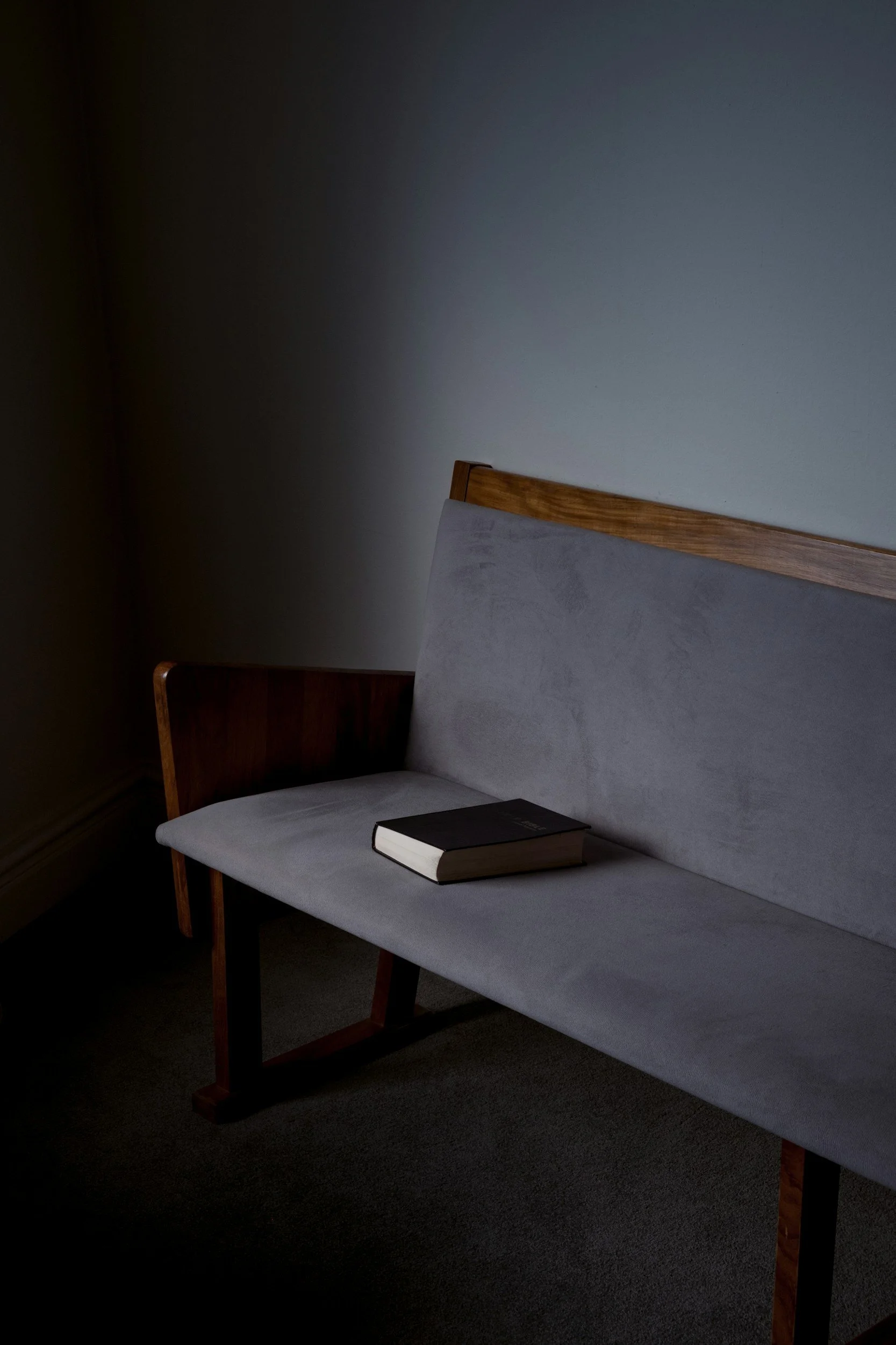 A gray upholstered bench with wooden legs and armrest, with a closed black book resting on it, in a dimly lit room with plain white walls.