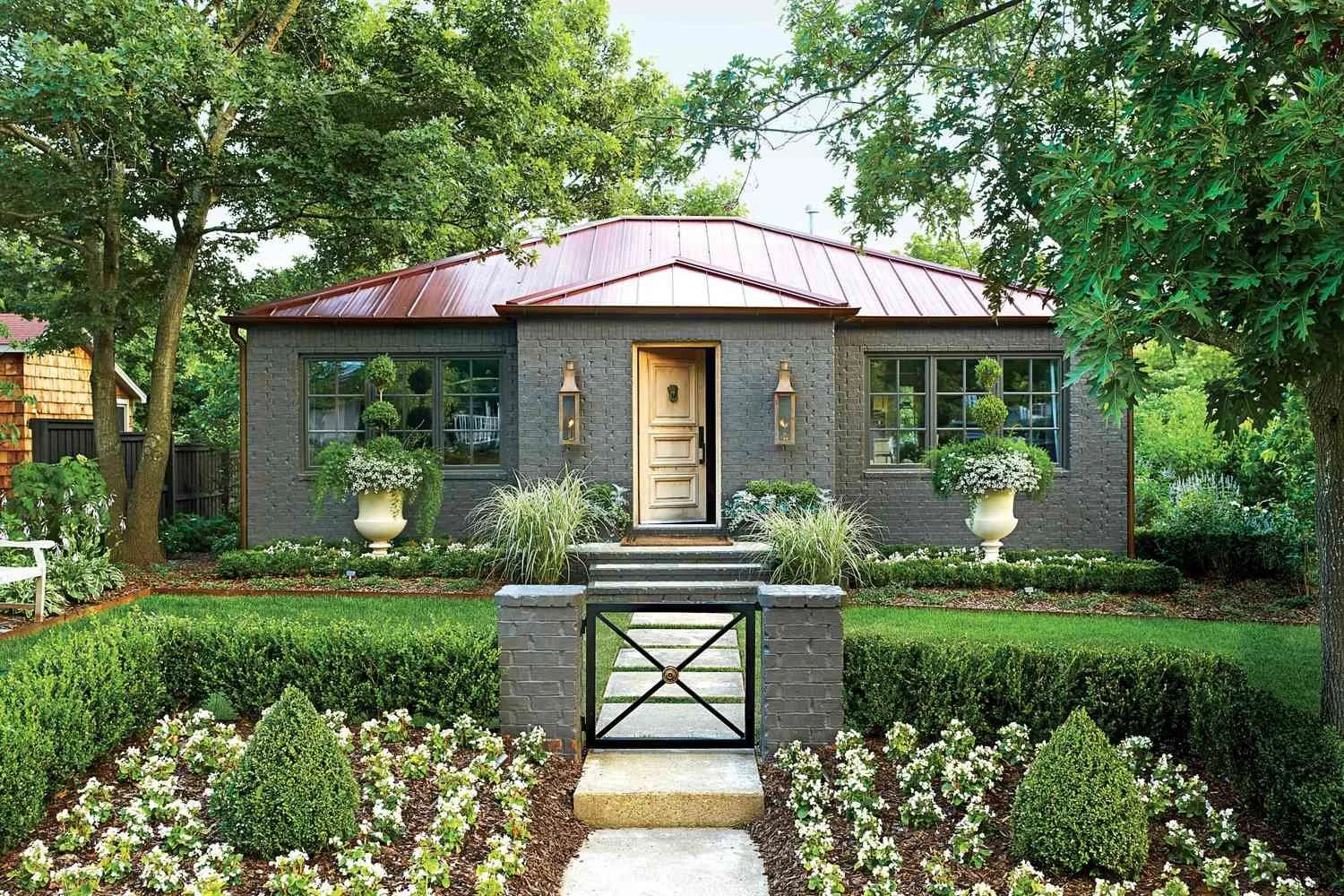 Front view of a small house with a red metal roof, dark brick walls, and a white front door. The house has large windows with plants and flowers on the window sills. There are two large potted plants with white flowers on either side of the door, and decorative lanterns mounted on the wall. A short brick and metal gate leads to the front steps, which are surrounded by a well-maintained garden with green bushes, flowering plants, and trees.