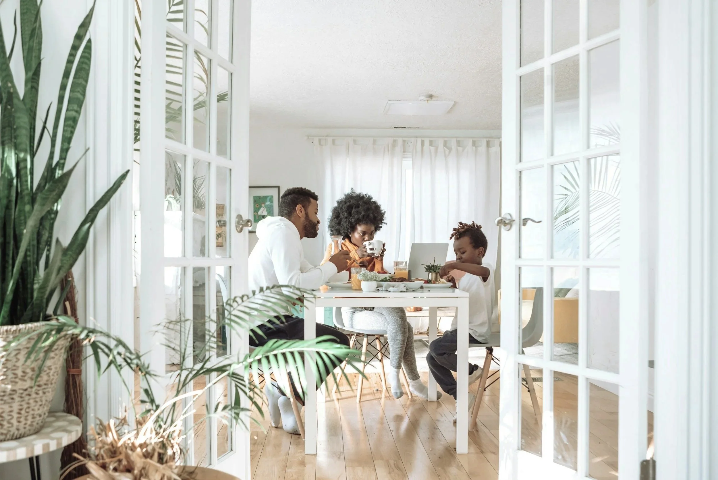 A family of four having breakfast together at a white dining table in a bright room with white curtains and wooden flooring, viewed through an open glass door with white frames.