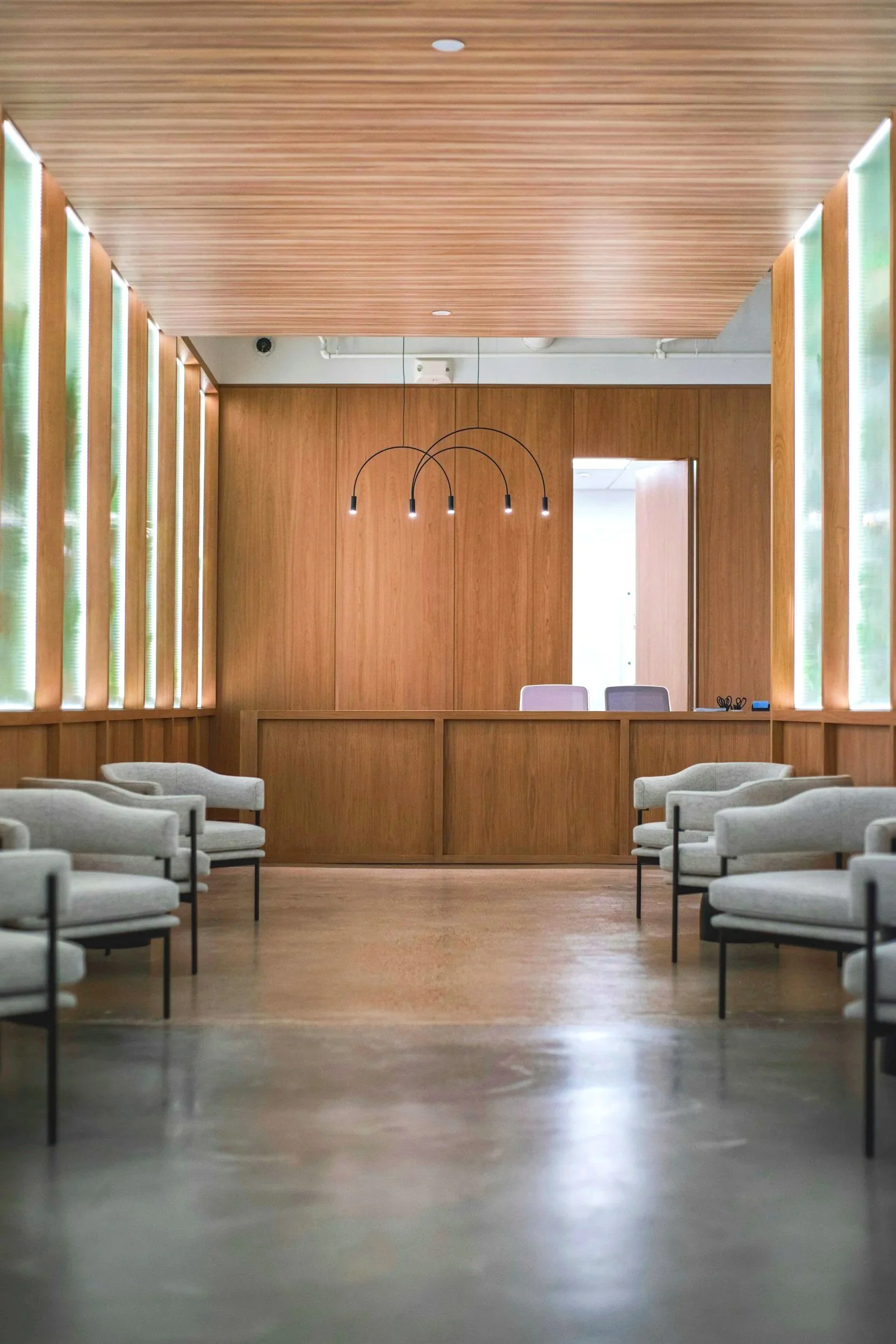 Empty waiting room with light gray chairs arranged along the walls, a wooden reception desk, and a ceiling with wood paneling. Modern black pendant lights hang above the desk, and there is a door open behind the desk leading to another room.