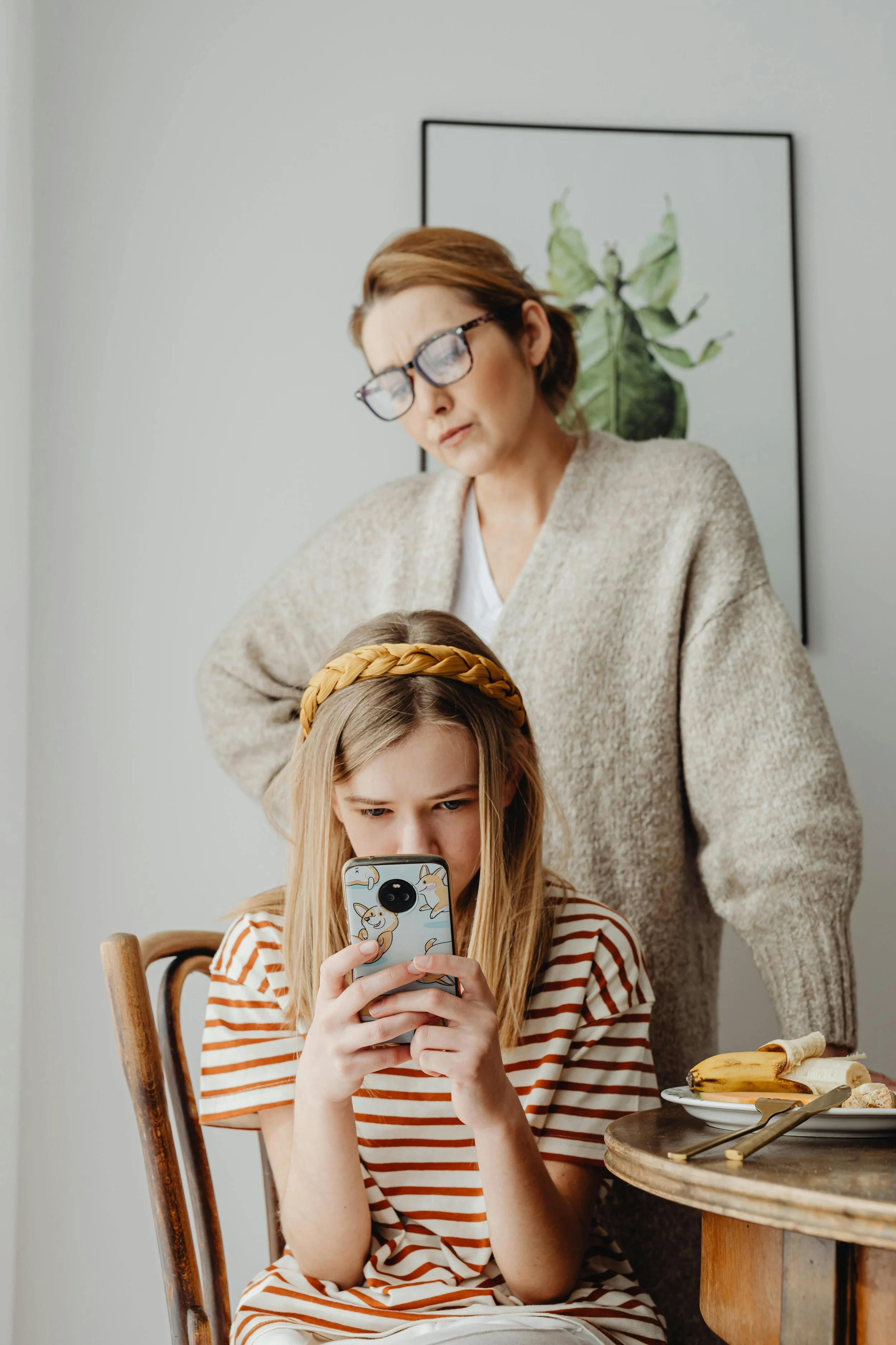 A woman with glasses and a beige sweater looks confused or concerned as a young girl with a braid and headband uses a smartphone at a table with bananas and bread.