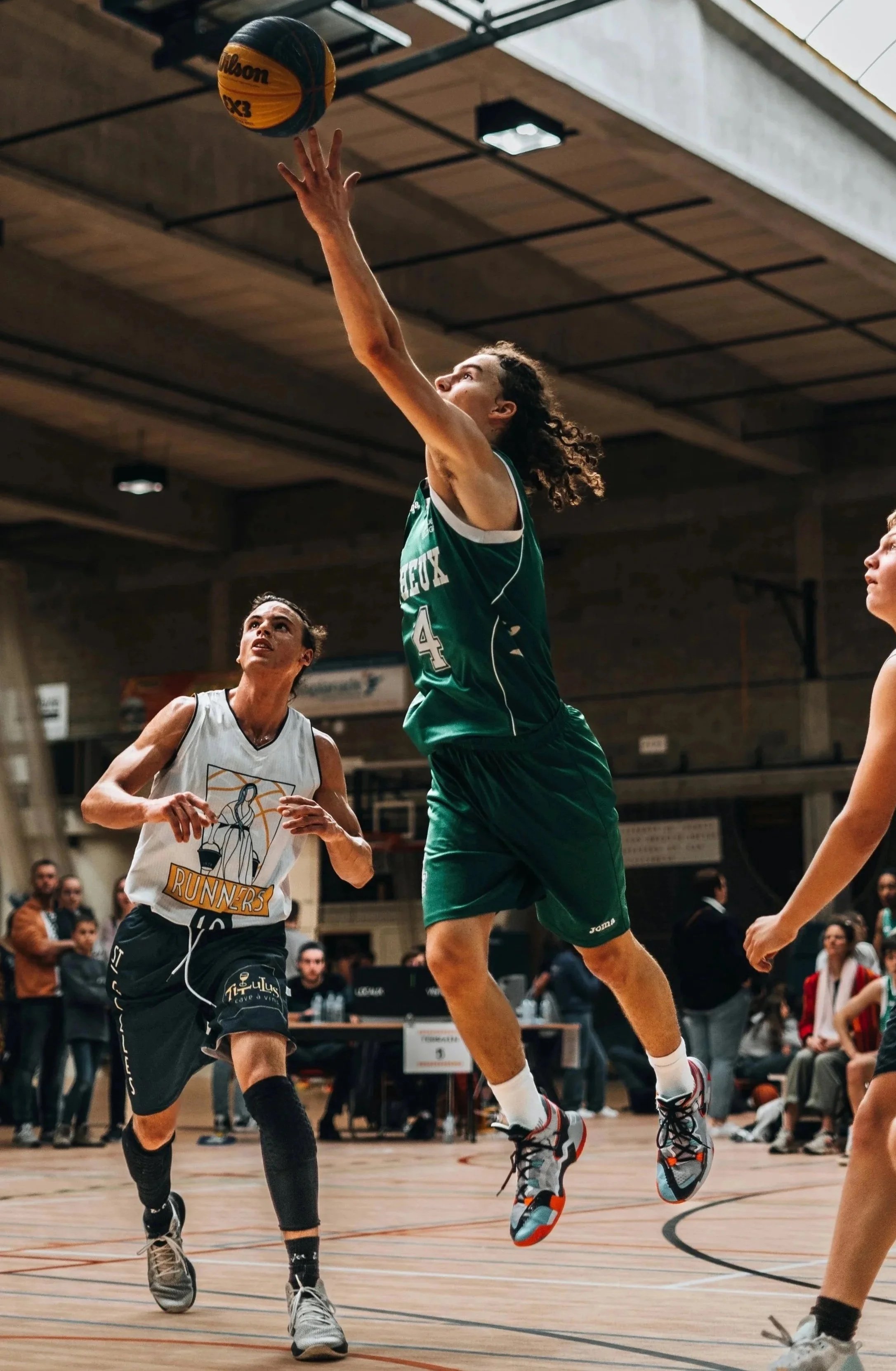 A female basketball player in a green uniform jumps to shoot or block the ball during a game, with two other players and spectators in the background.