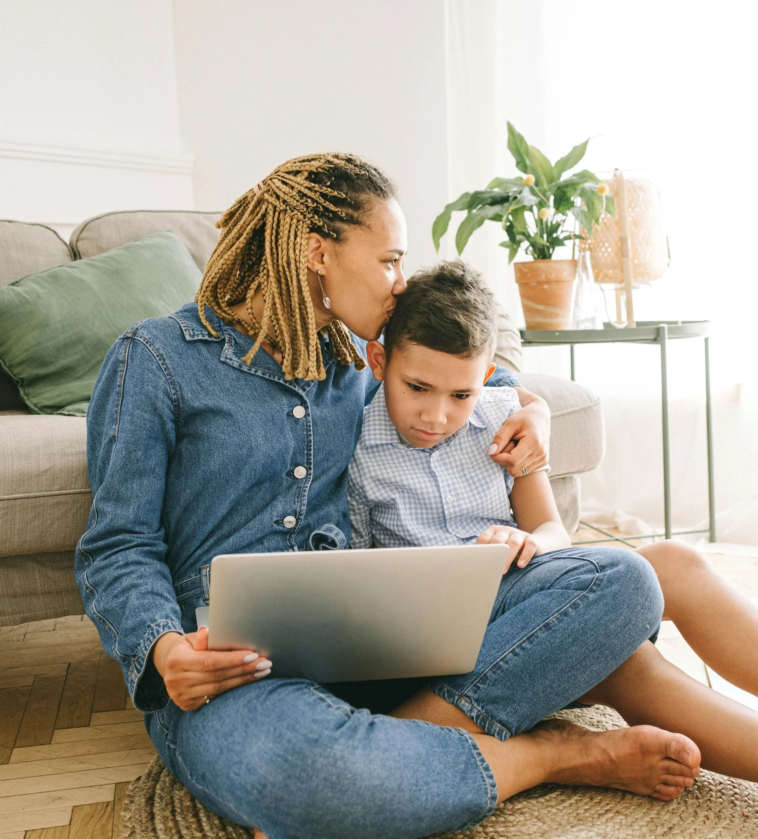 A woman with braids and a denim jacket sitting on the floor, kissing a young boy on the head while they look at a laptop together in a living room with a sofa, green pillow, potted plant, and lamp in the background.