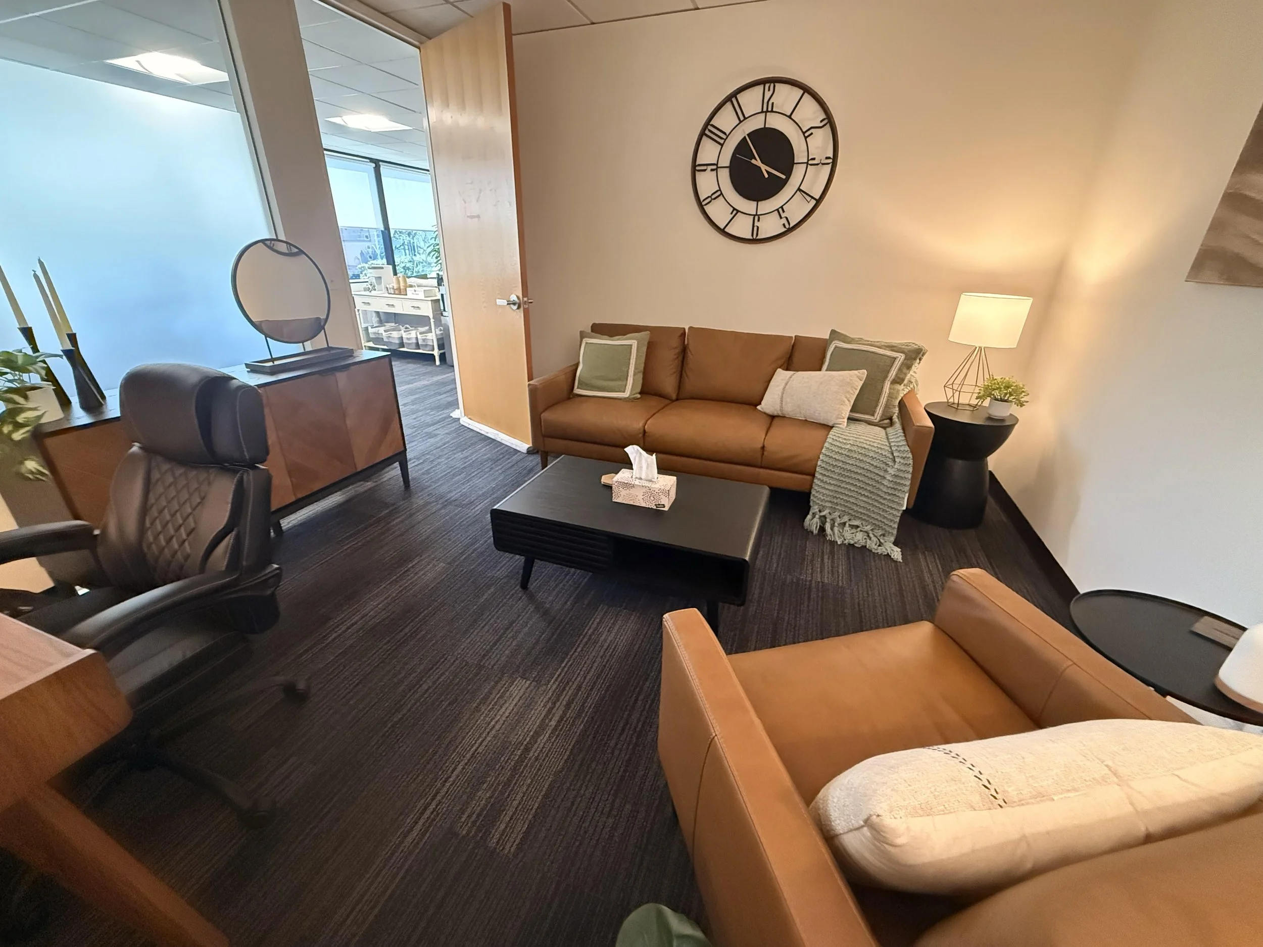 Waiting room with brown leather sofa, orange armchair, black coffee table, side table with lamp, and decorative pillows, with a wall clock above the sofa.