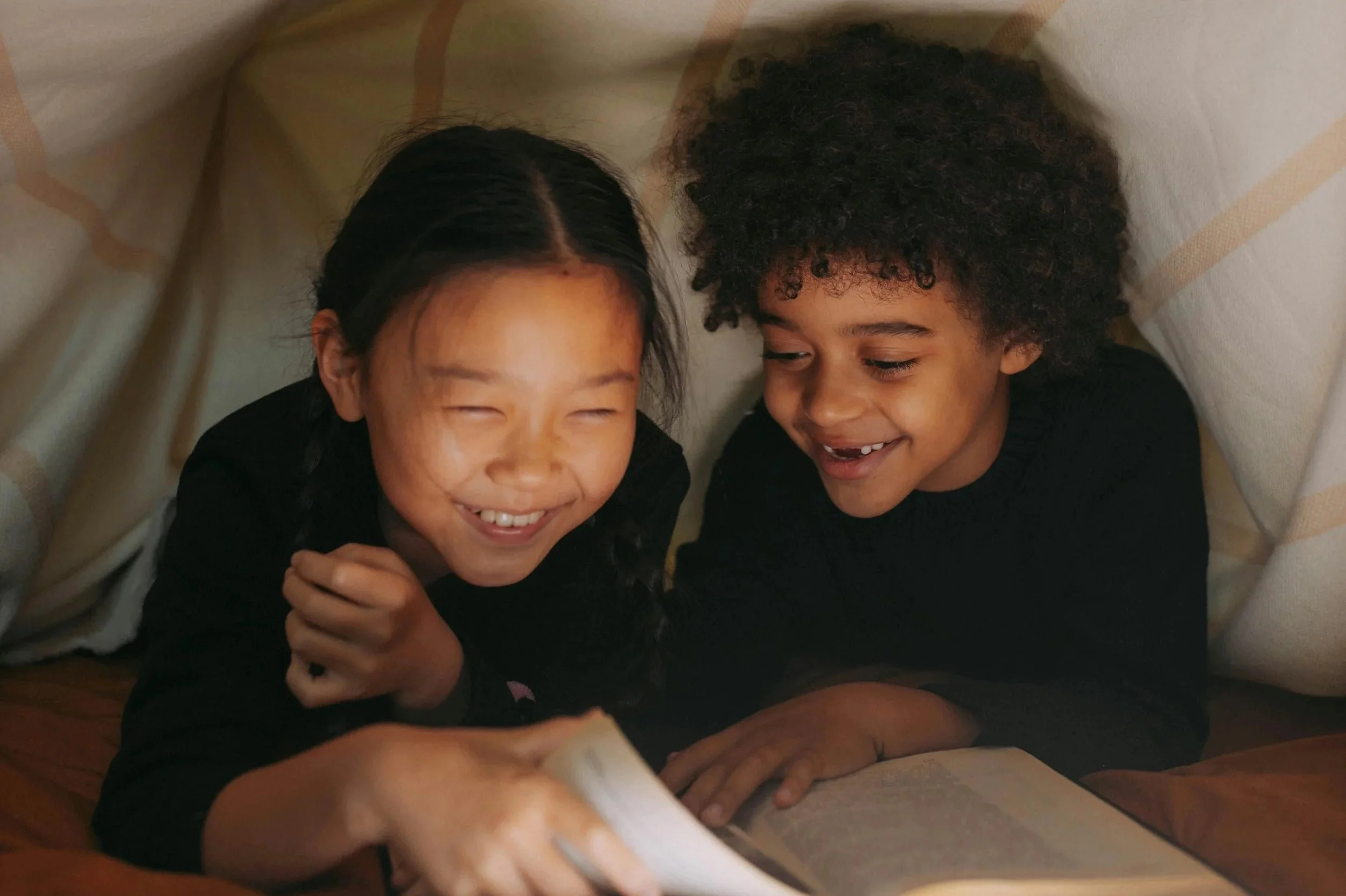 Two children, a girl with braided hair and a boy with curly hair, smiling and looking at a book inside a makeshift fort made of blankets.