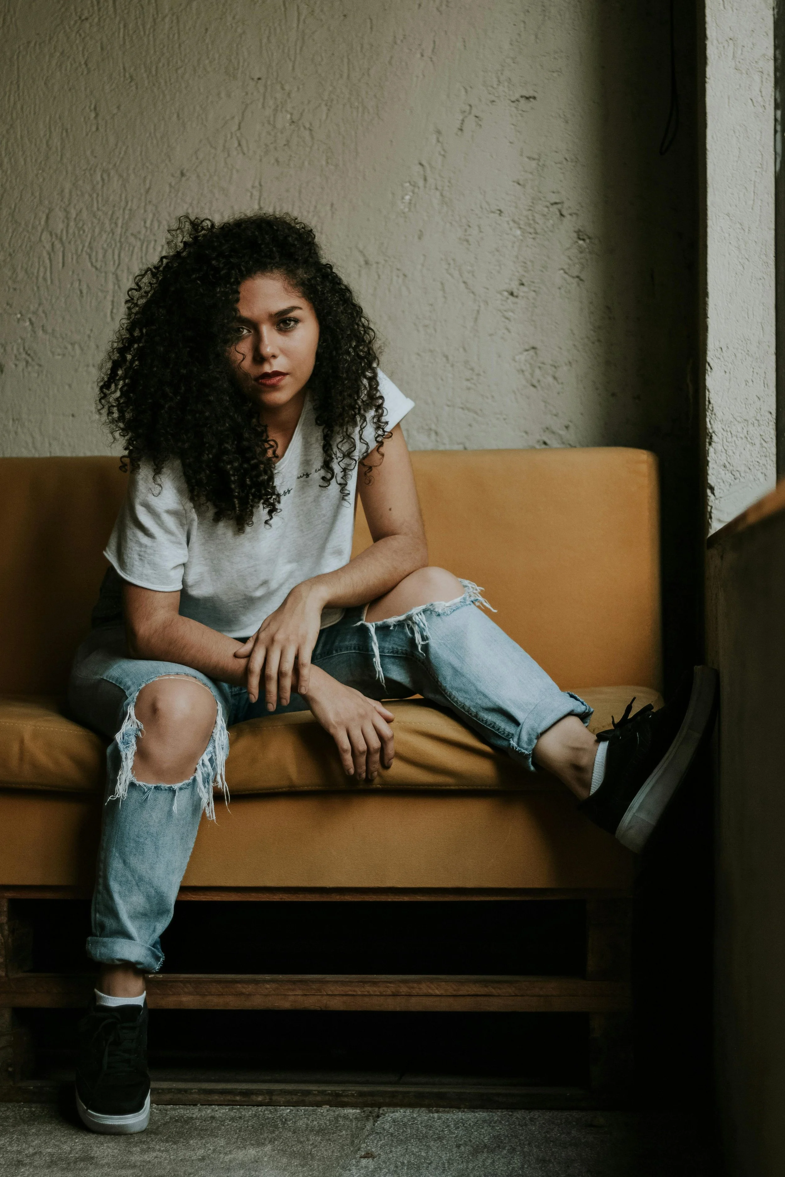 A young woman with curly hair sitting on a tan couch, wearing a white T-shirt, ripped jeans, and black sneakers, in a room with textured beige walls.