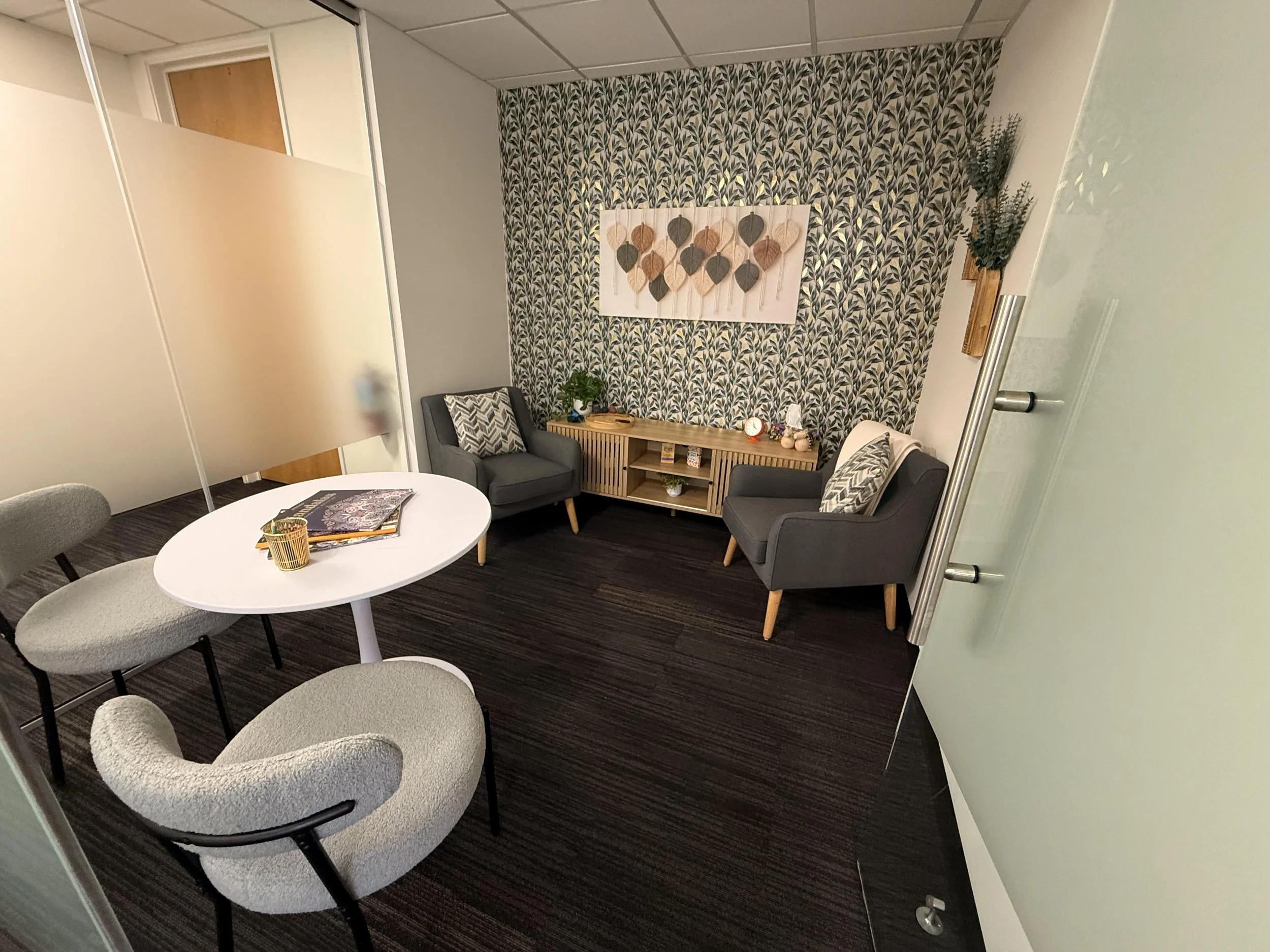 Office with two gray armchairs, patterned pillows, a small wooden side table, a decorative wall hanging, a patterned wallpaper accent wall, a white round table with magazines, and a whiteboard or frosted glass partition.