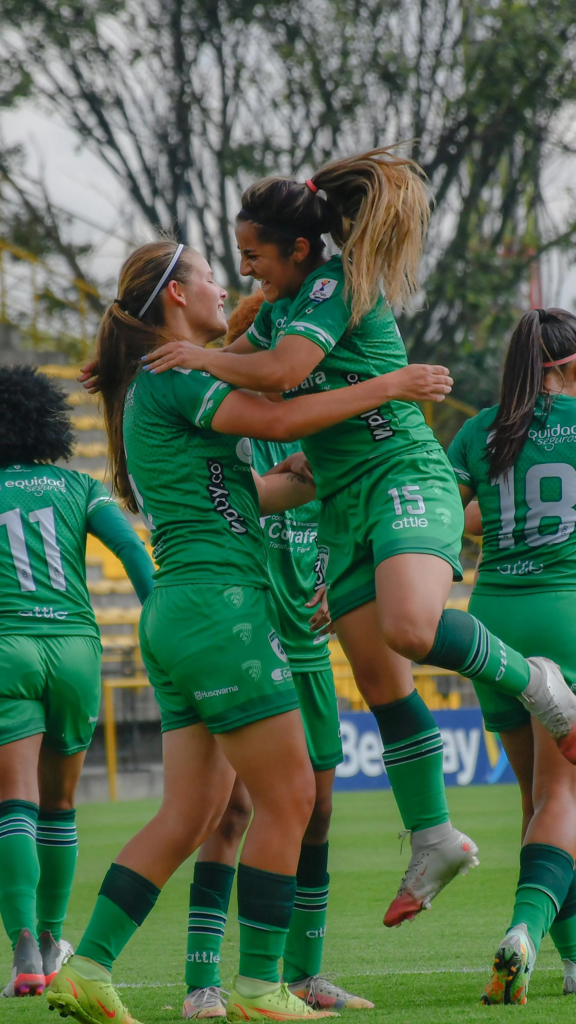 Two female soccer players in green uniforms celebrating on the field. One player is jumping into the arms of the other, and they are smiling and holding each other.