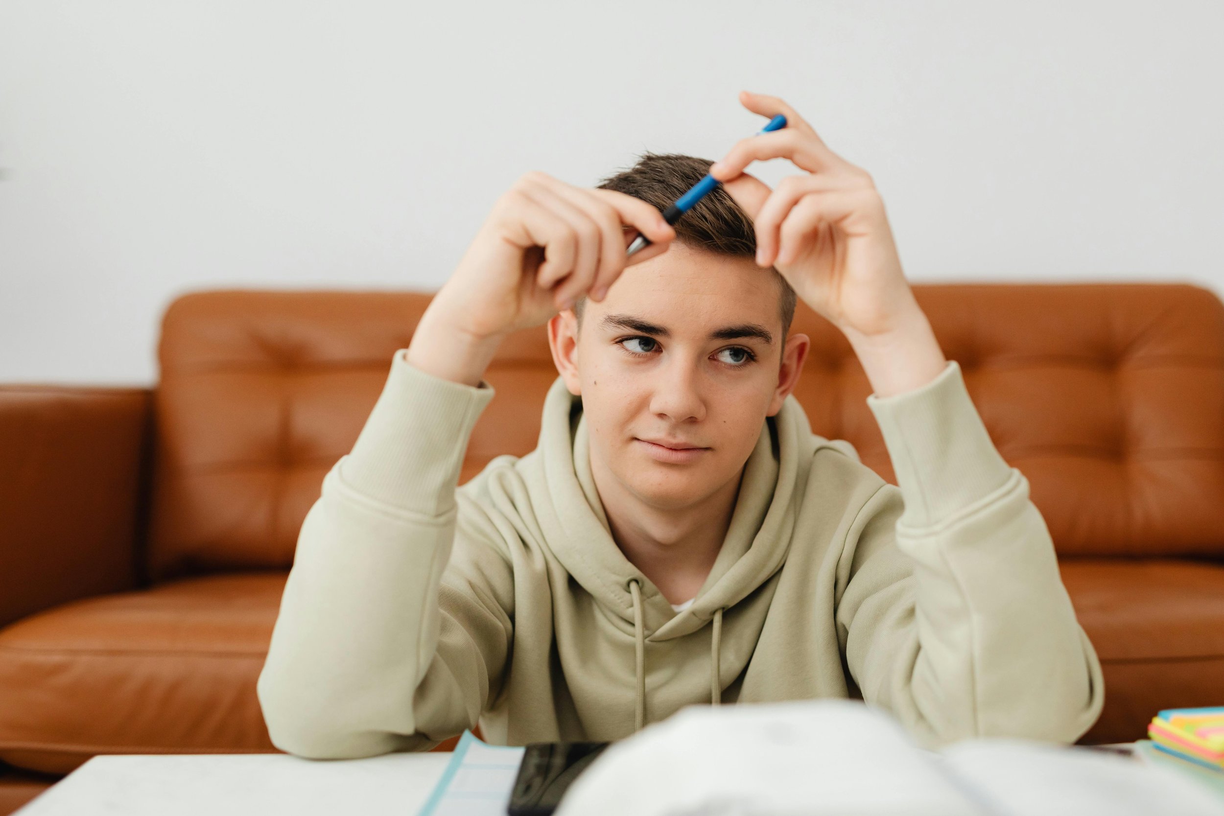 Teenager boy sitting at a table, holding a pen above his head, looking to the side with a neutral expression, with books and notebooks on the table and a brown couch in the background.