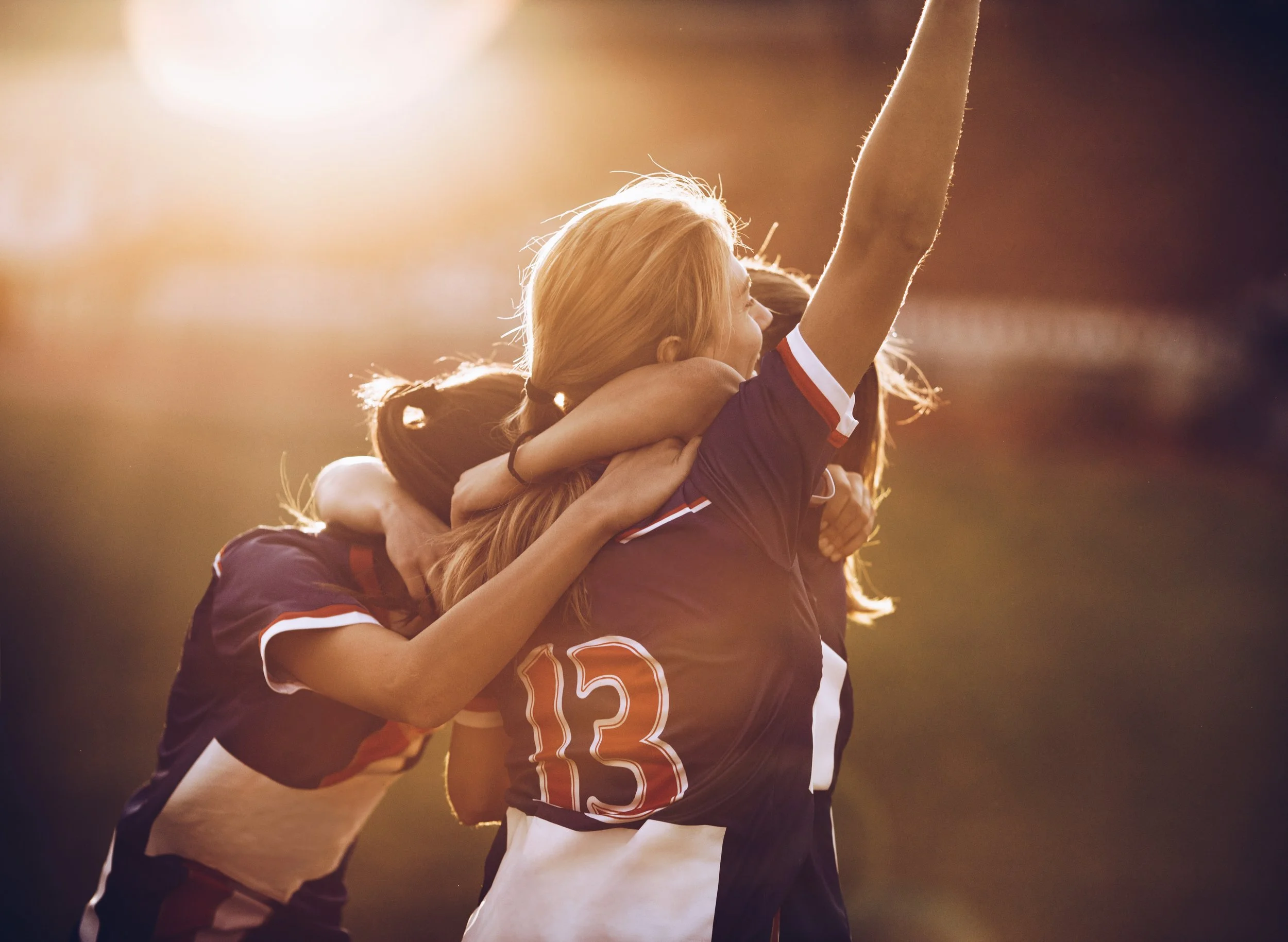 Group of female athletes celebrating on the field during sunset, with one woman raising her arm in victory.