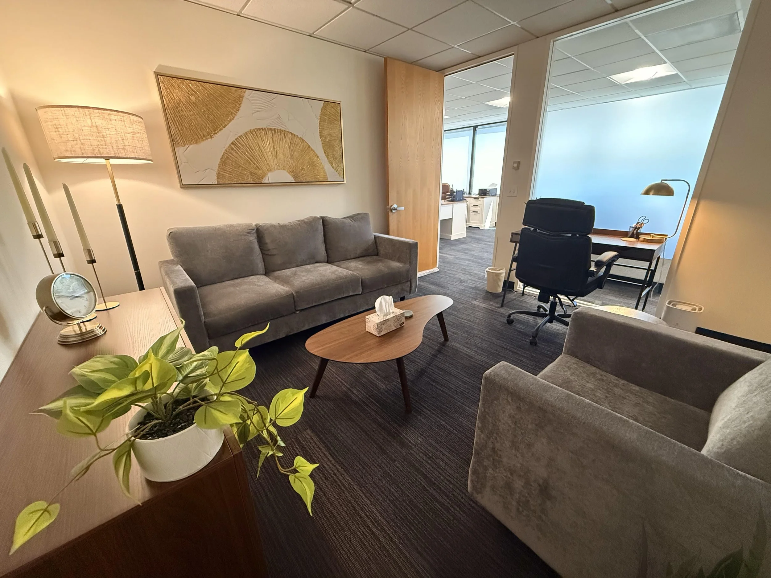 Office waiting area with two gray sofas, a wooden coffee table with tissue box, a potted plant on sideboard, wall art, standing lamp, and a desk with an office chair, all in a modern, well-lit space.