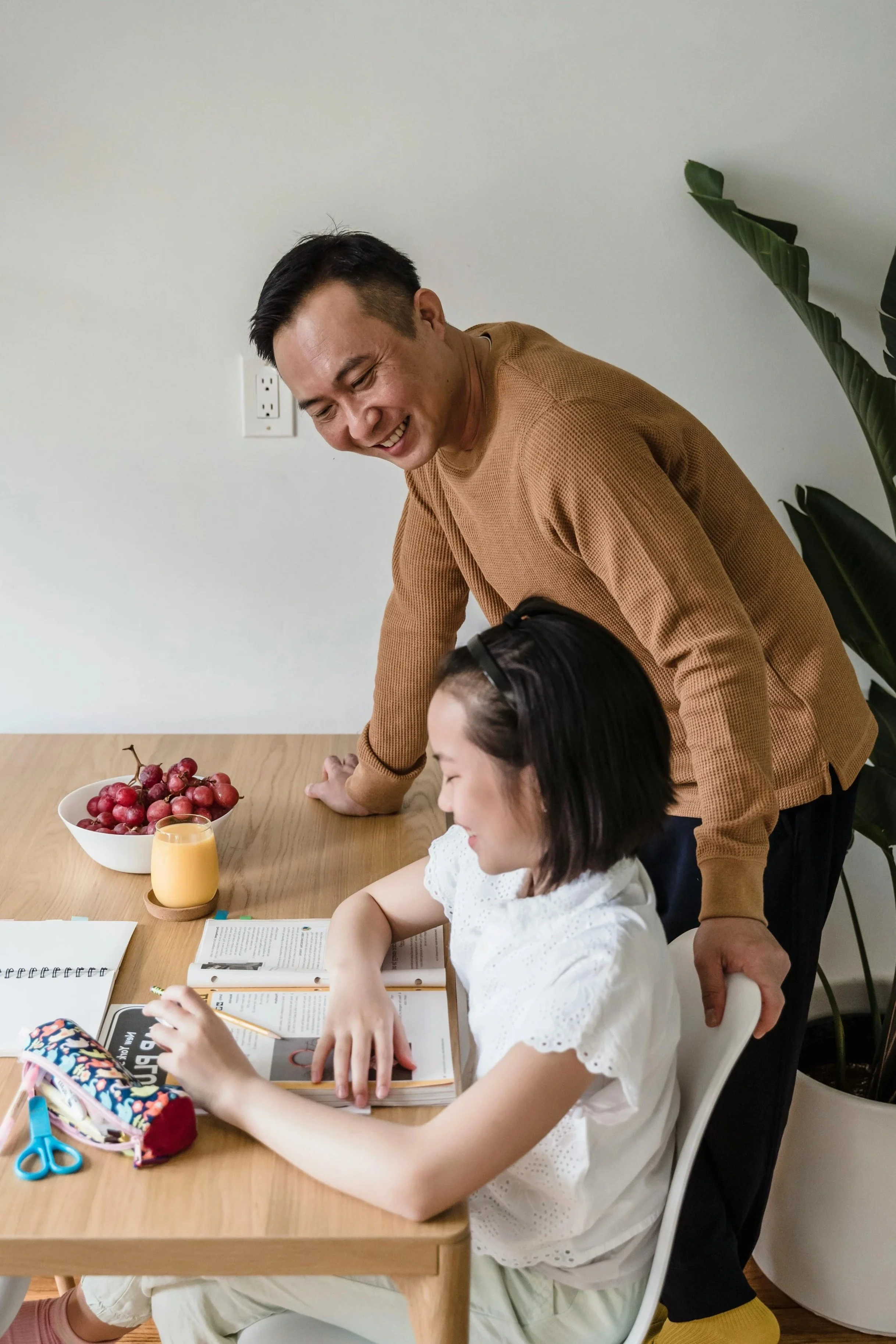 A young girl sitting at a wooden table, smiling, with an open book and a pencil in hand, while an adult man leaning over her, smiling and engaging in conversation. On the table, there are a bowl of grapes, a glass of orange juice, colorful scissors, a pencil case, and notebooks.