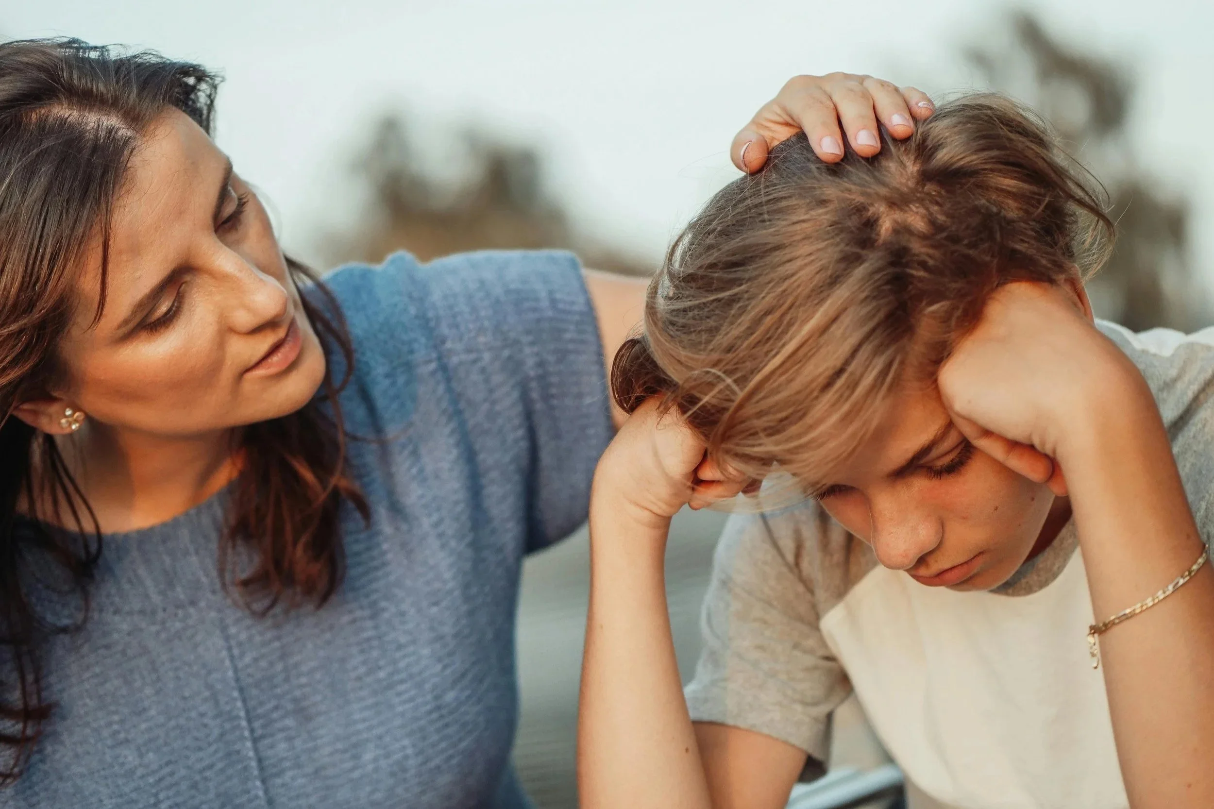A woman comforts a distressed teenage boy who is holding his head and looking down.