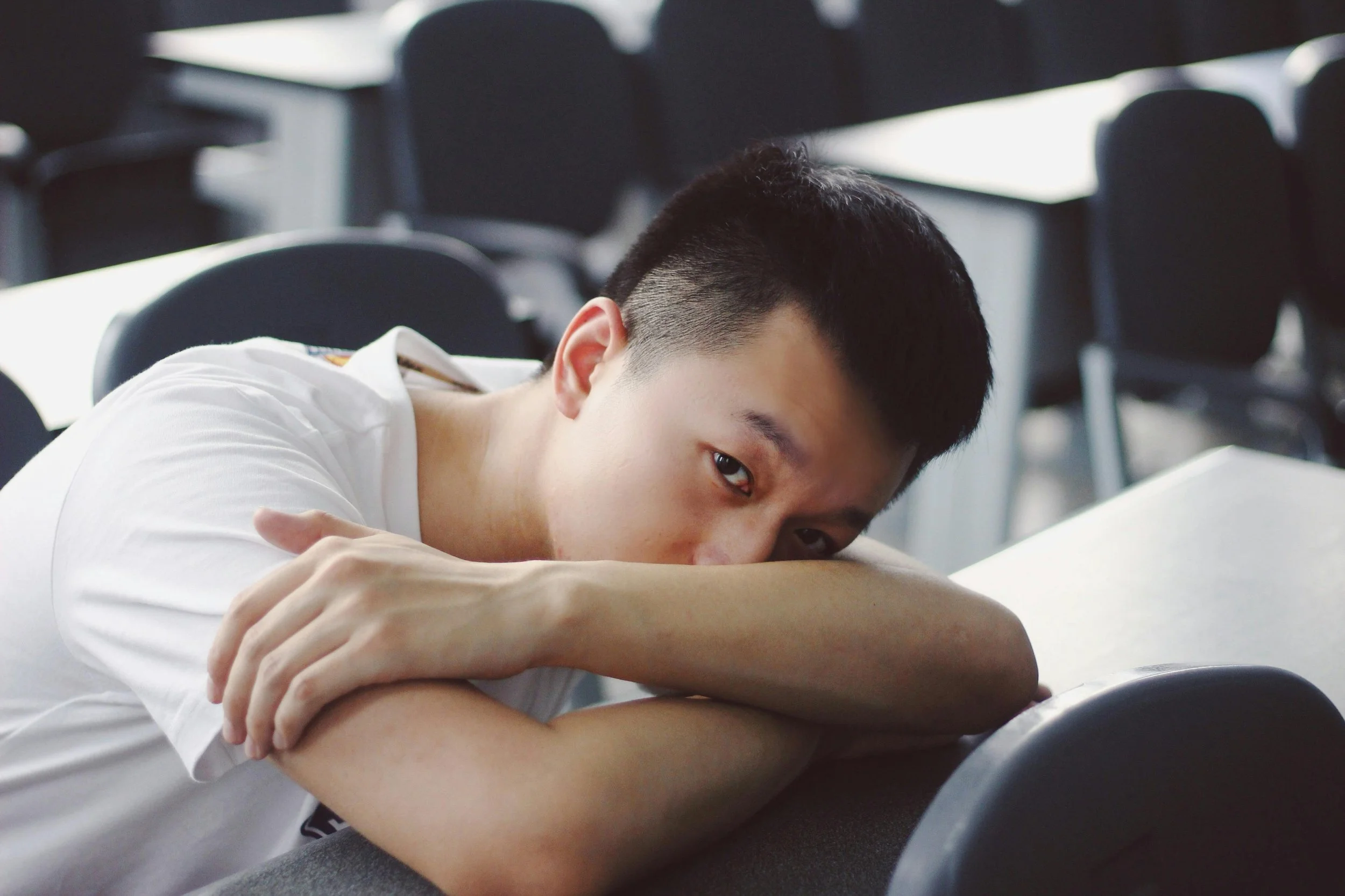 A young person with short black hair resting their head on their arms, sitting at a table in a room with black chairs and tables.