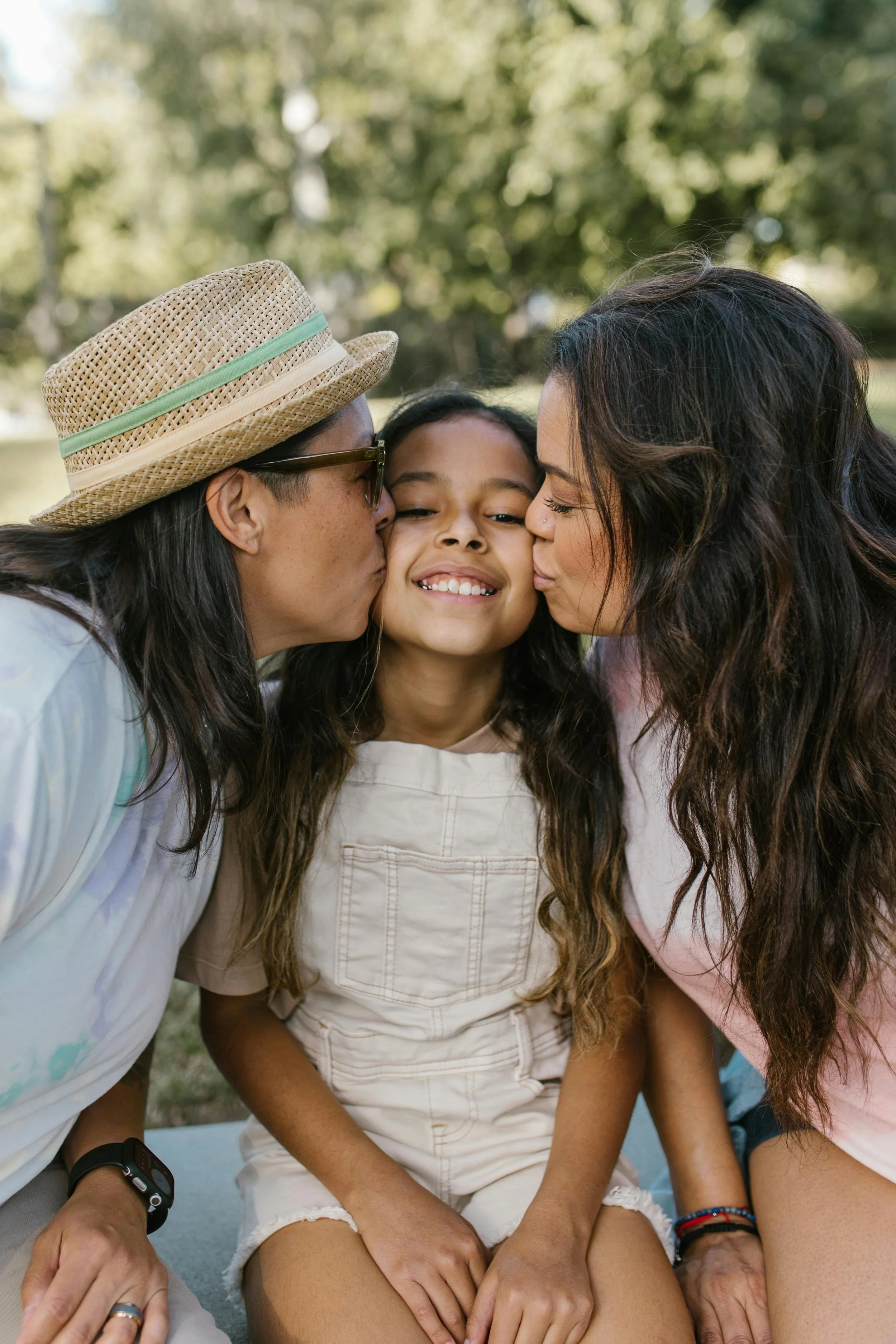 A young girl sitting outdoors as her parents kiss her on the cheeks during daytime with a blurred background of trees.