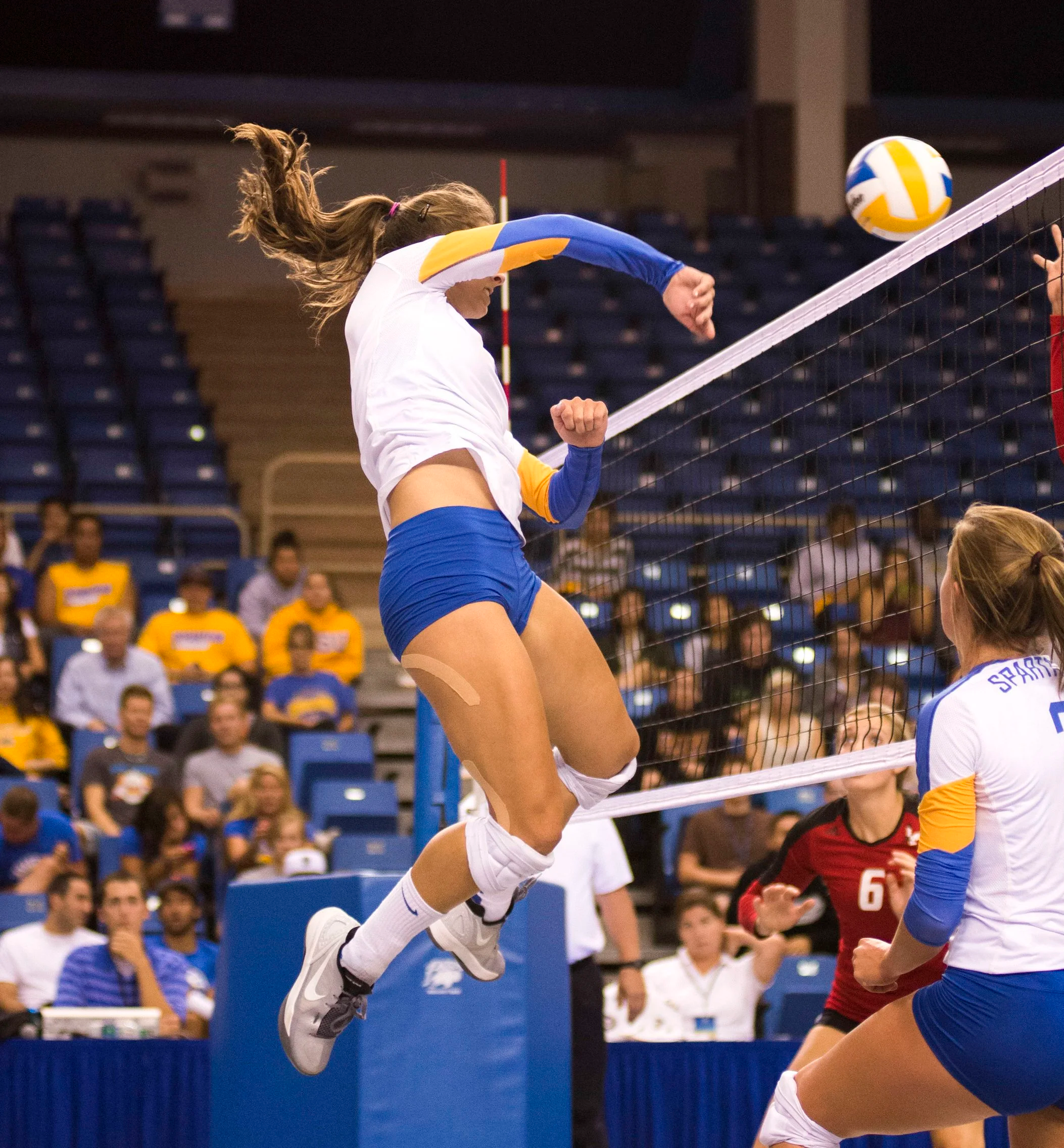 A female volleyball player jumping at the net during a match, with spectators in the background.
