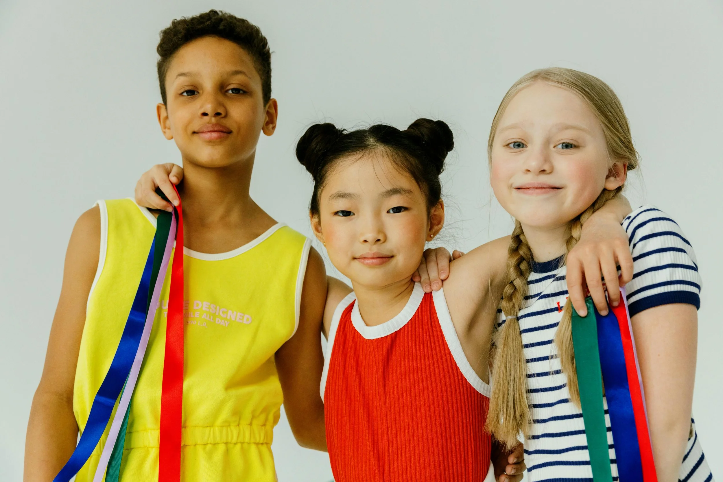 Three young girls stand together with their arms around each other, smiling, with colorful ribbons in their hands, against a plain white background.