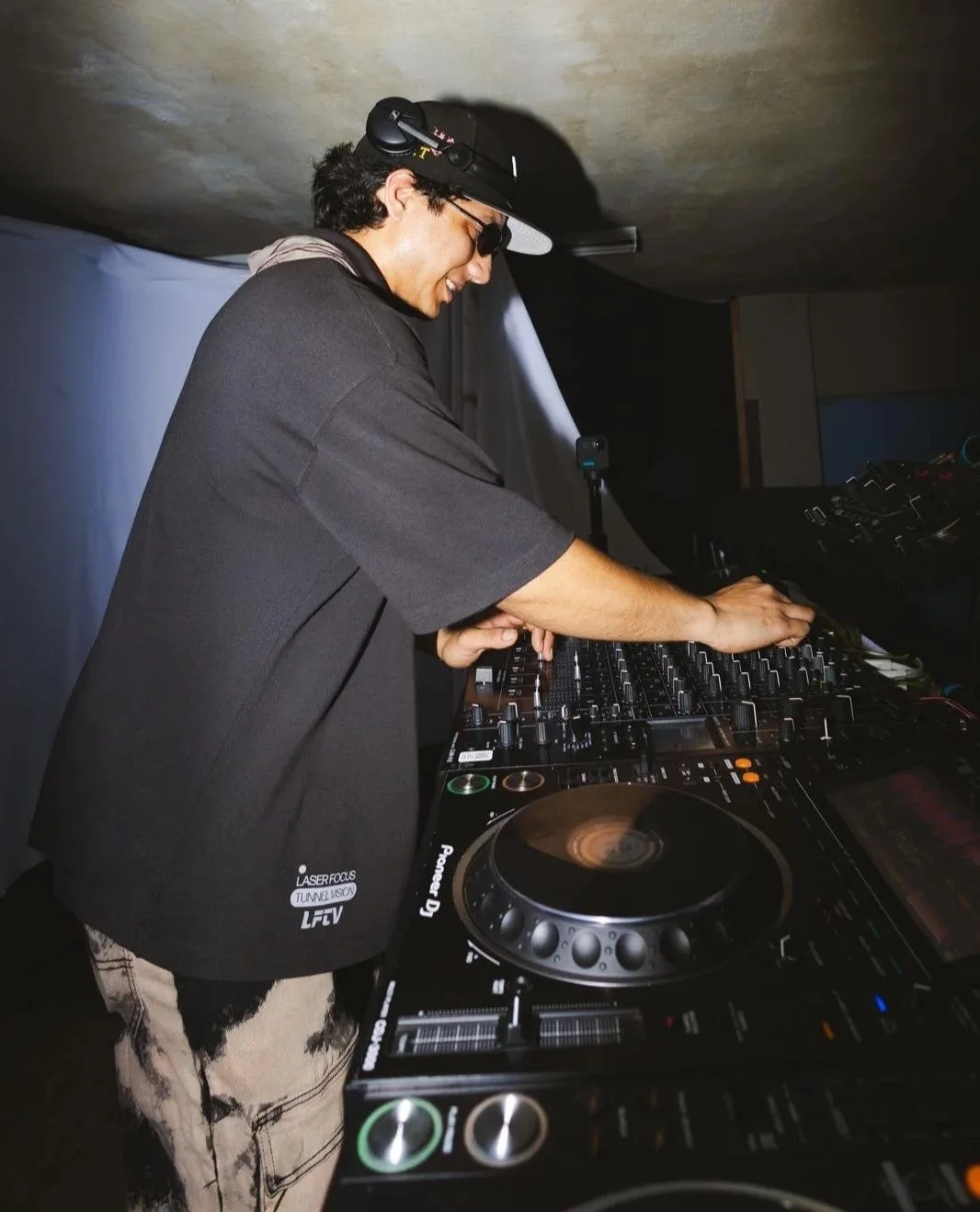 A DJ wearing sunglasses and a black cap is smiling while mixing music on a DJ controller in a dimly lit environment.