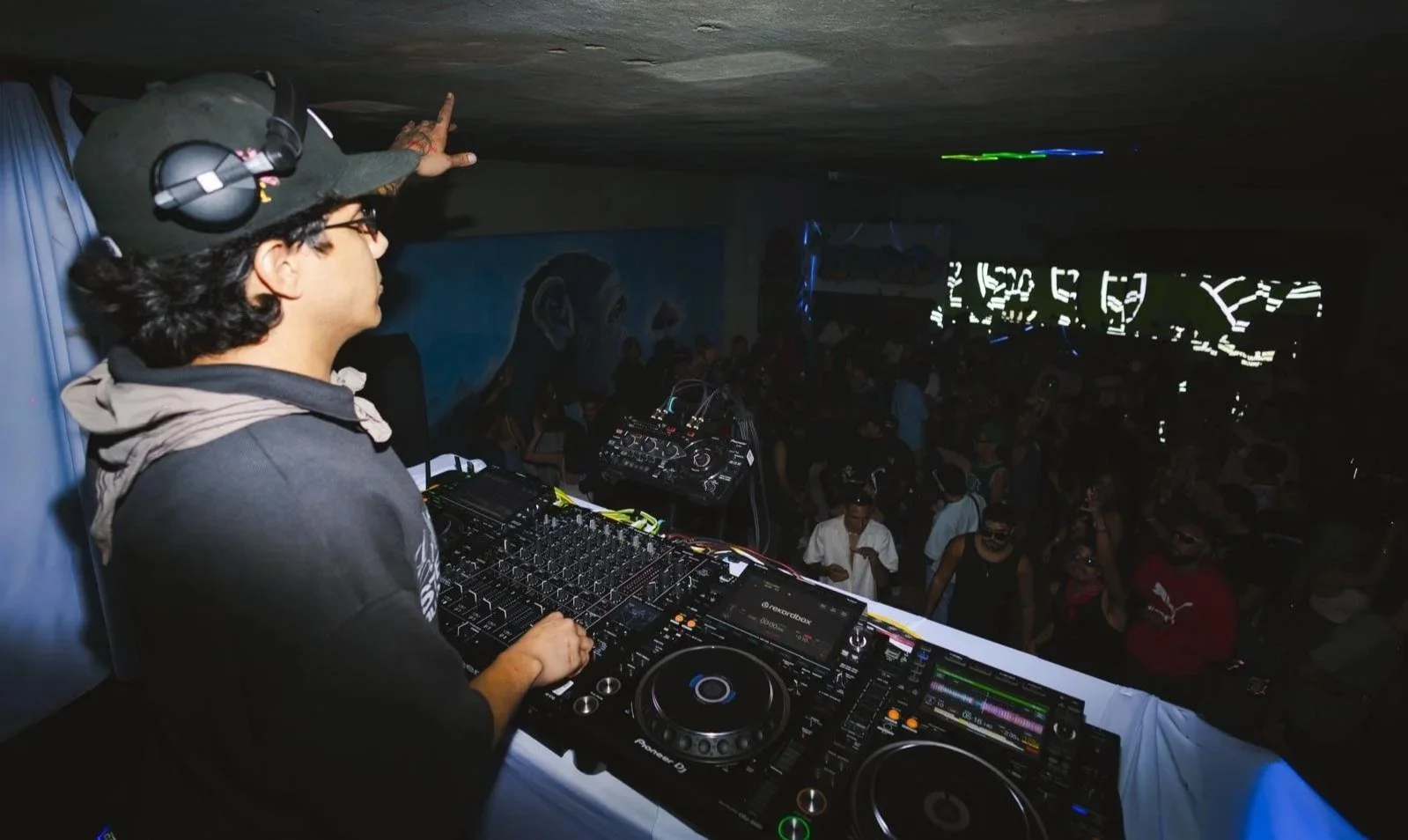 A DJ standing at a mixing console with headphones and microphone, performing at a crowded indoor event with many people dancing, colorful lights, and a mural on the wall in the background.