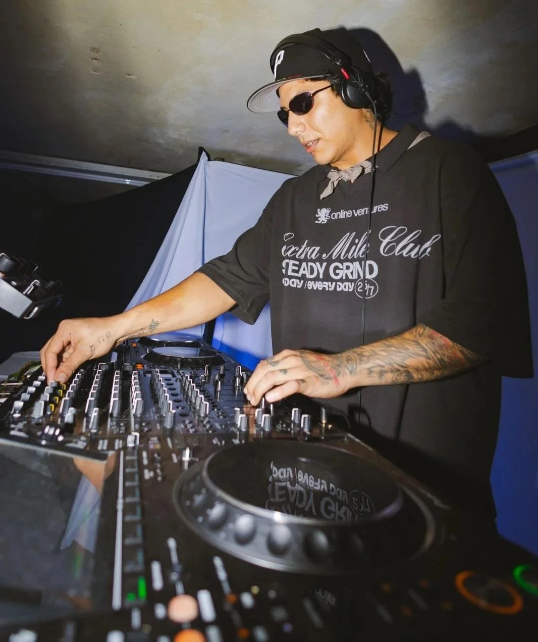 A DJ wearing sunglasses, a black cap, headphones, and a black T-shirt with white text, is mixing music on a DJ console.
