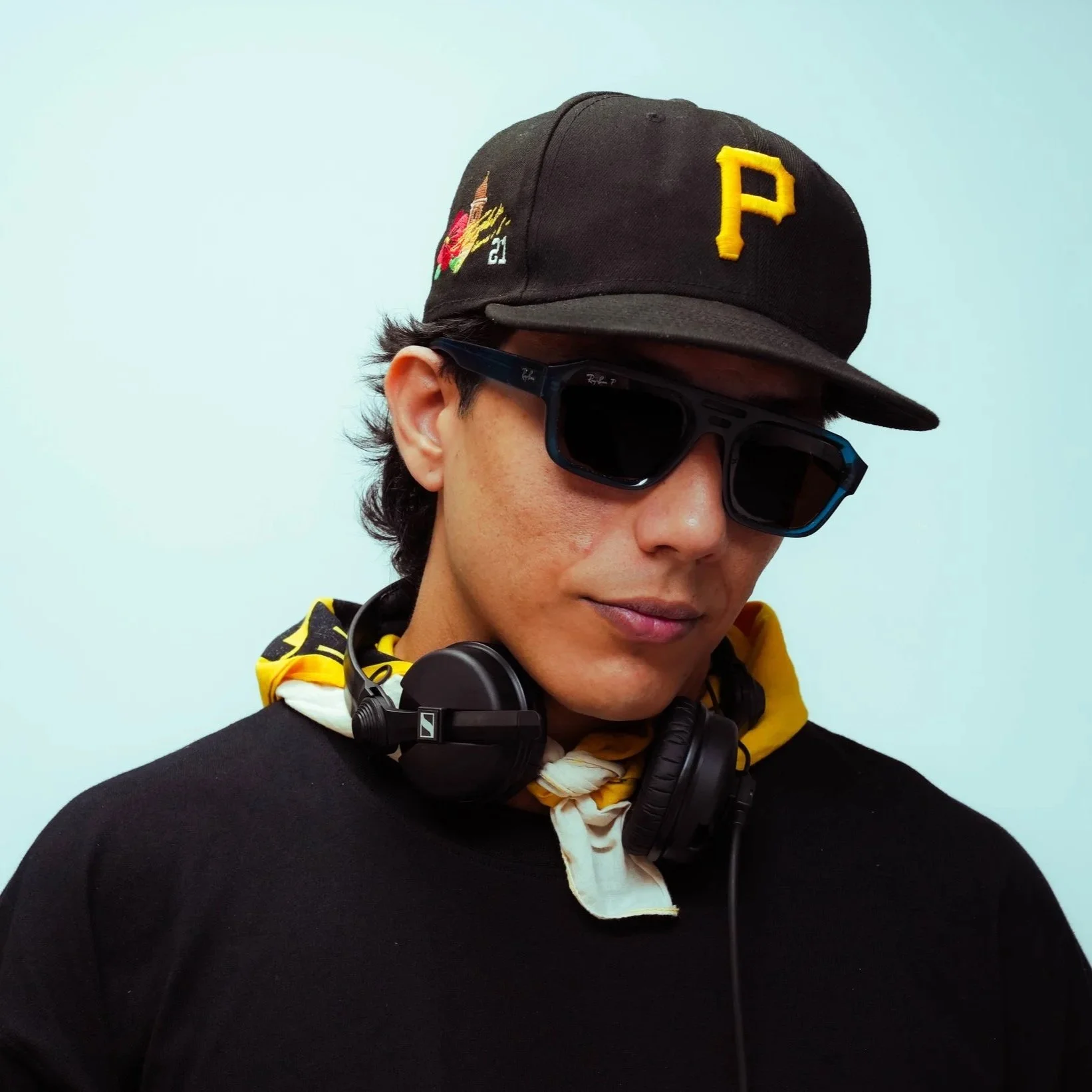 Young man wearing a black baseball cap with a yellow P, black sunglasses, and a black shirt, with headphones around his neck and a yellow bandana around his neck, against a light blue background.