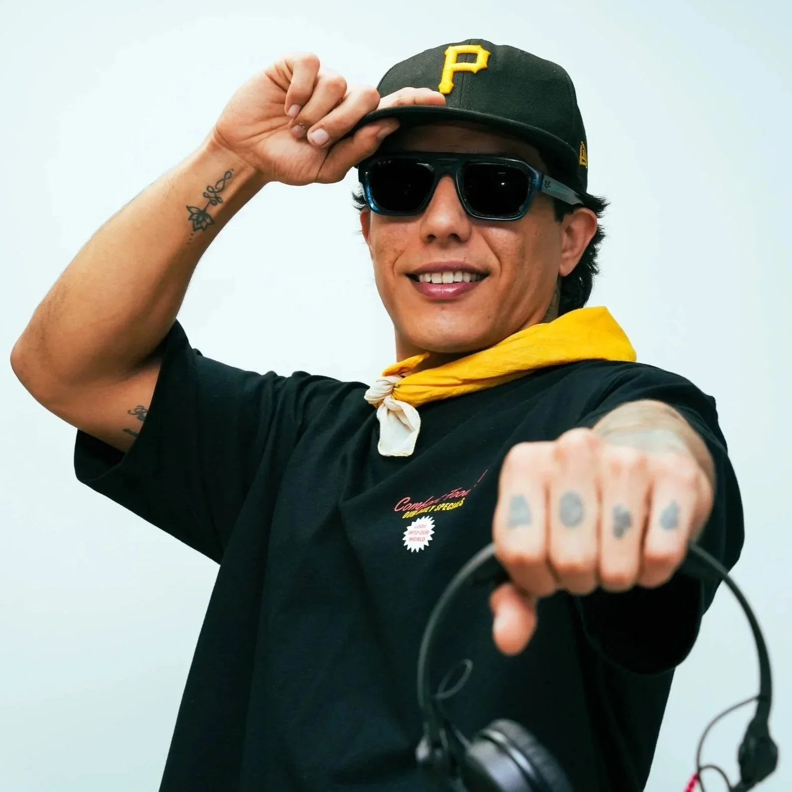 A man wearing sunglasses, a black baseball cap with a yellow 'P', a black T-shirt, and a yellow bandana around his neck is smiling and pointing towards the camera. He has tattoos on his arm and hand, and is holding headphones.