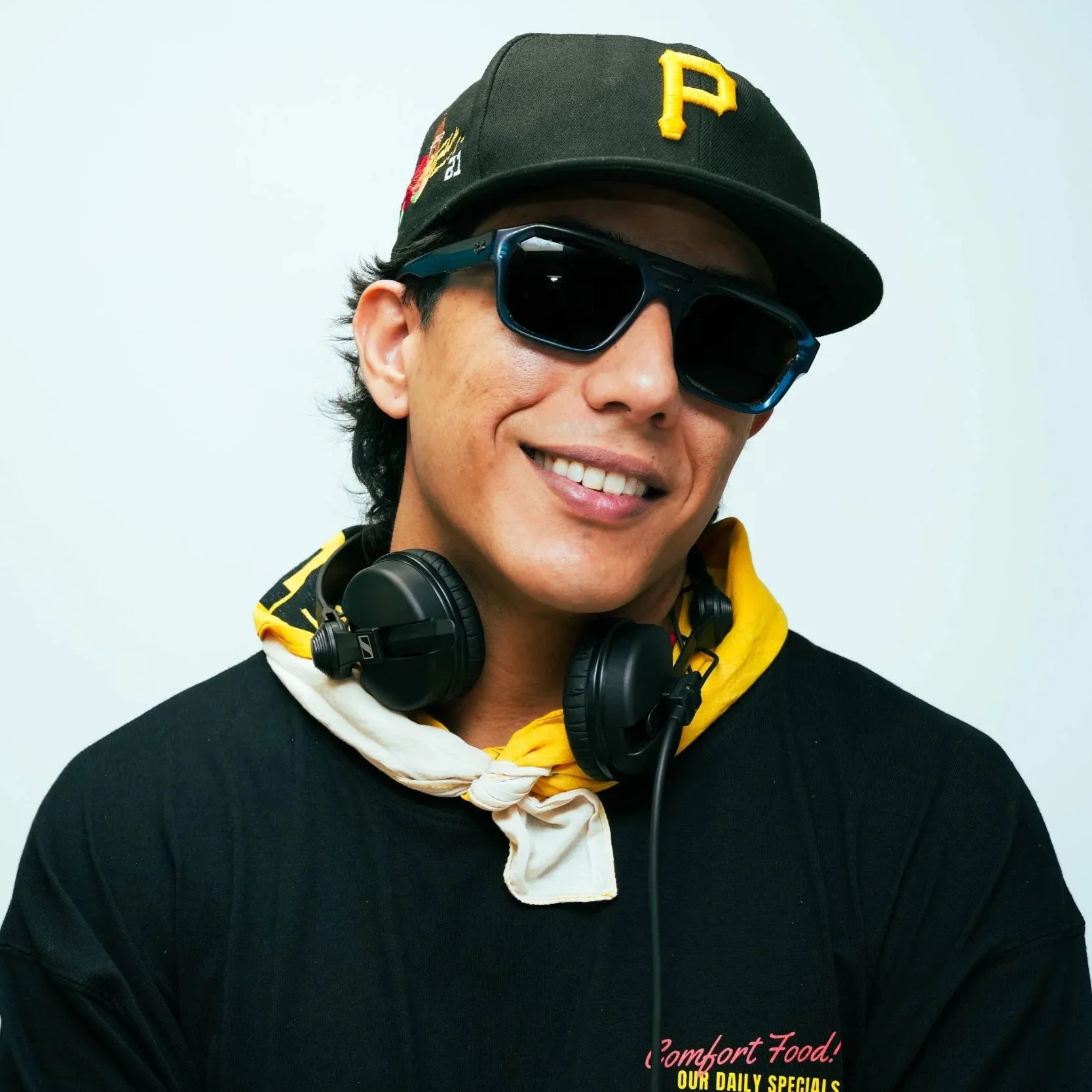 A smiling man wearing sunglasses, a black cap with a yellow 'P', a black T-shirt, a white and yellow neck scarf, and headphones around his neck.