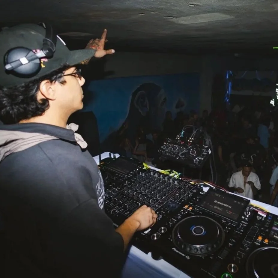 A DJ standing at a mixing console with headphones and microphone, performing at a crowded indoor event with many people dancing, colorful lights, and a mural on the wall in the background.