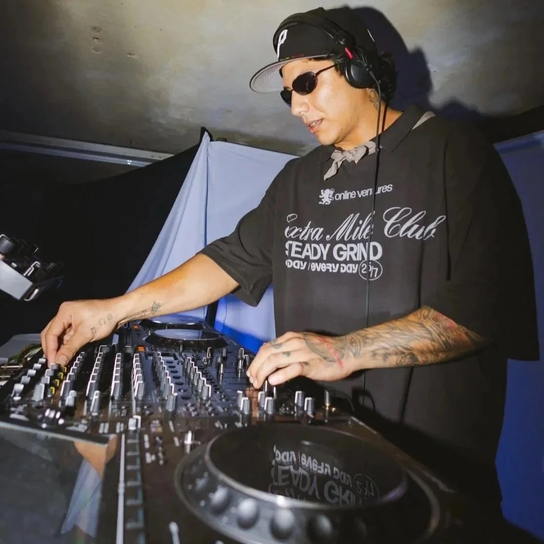 A DJ wearing sunglasses, a black cap, headphones, and a black T-shirt with white text, is mixing music on a DJ console.