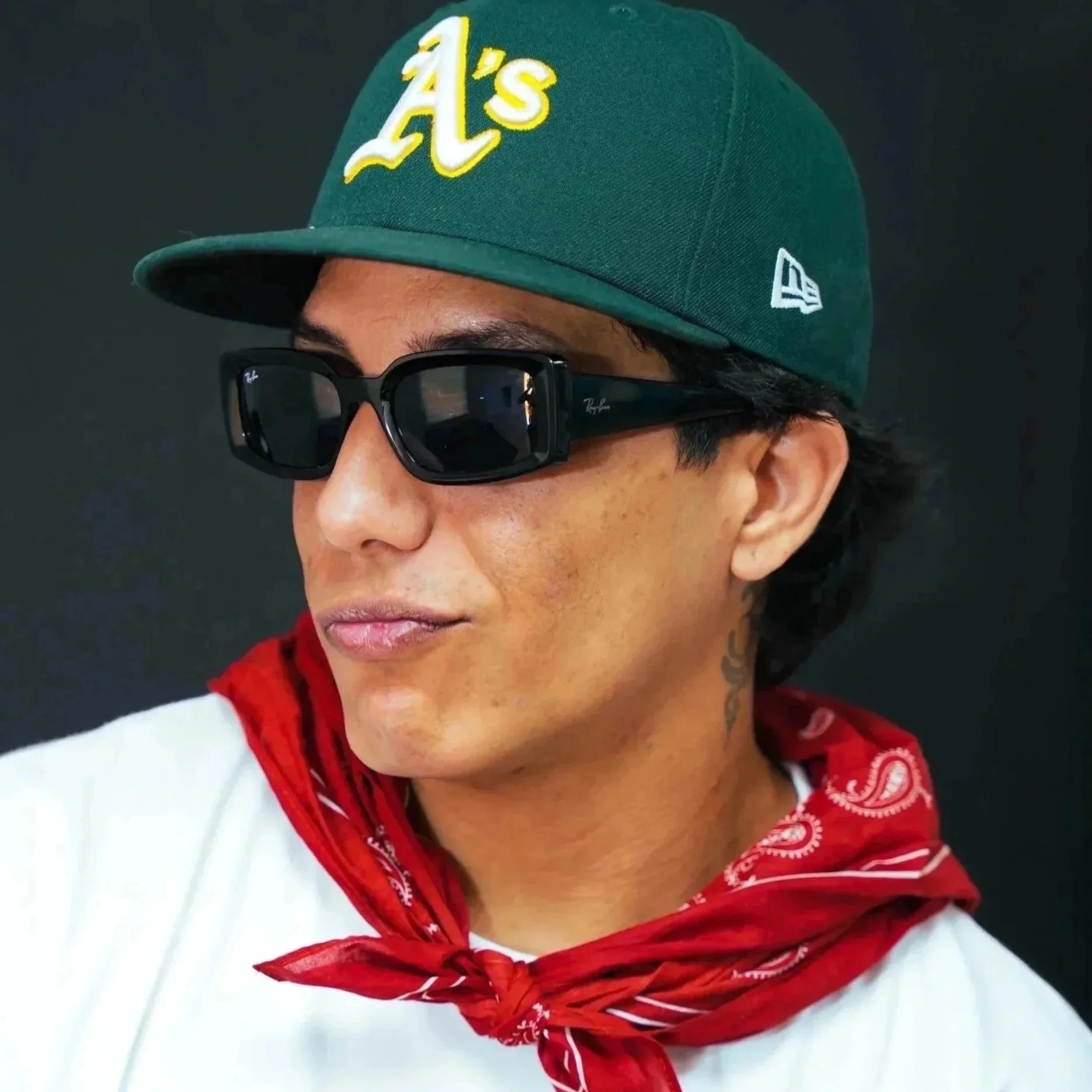A young man wearing a green Oakland Athletics baseball cap, black sunglasses, a red bandana around his neck, and a white T-shirt with a flower graphic and the words 'BLOOMFREE' printed on it.