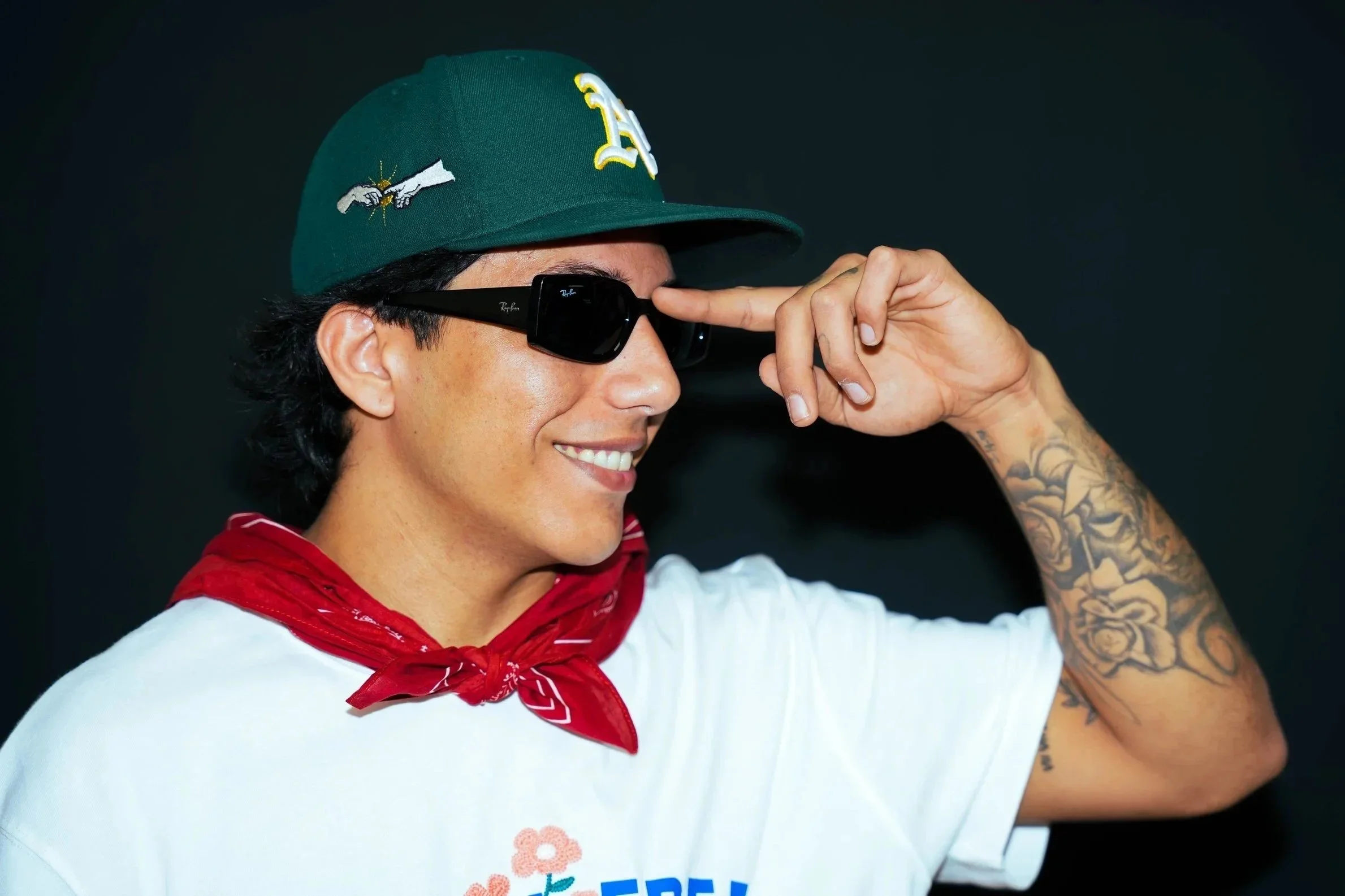 A person wearing a green baseball cap with embroidered patches, black sunglasses, a white T-shirt with logos, and a red bandana around their neck. They are smiling and saluting with their right hand, which has tattoos on the arm.