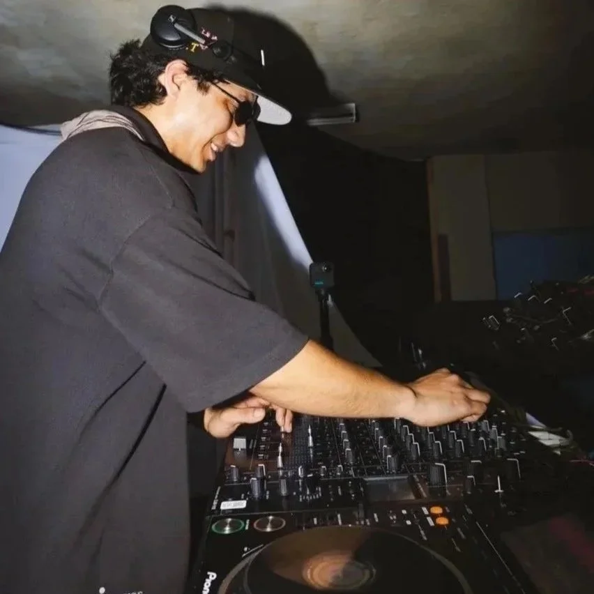 A DJ wearing sunglasses and a black cap is smiling while mixing music on a DJ controller in a dimly lit environment.
