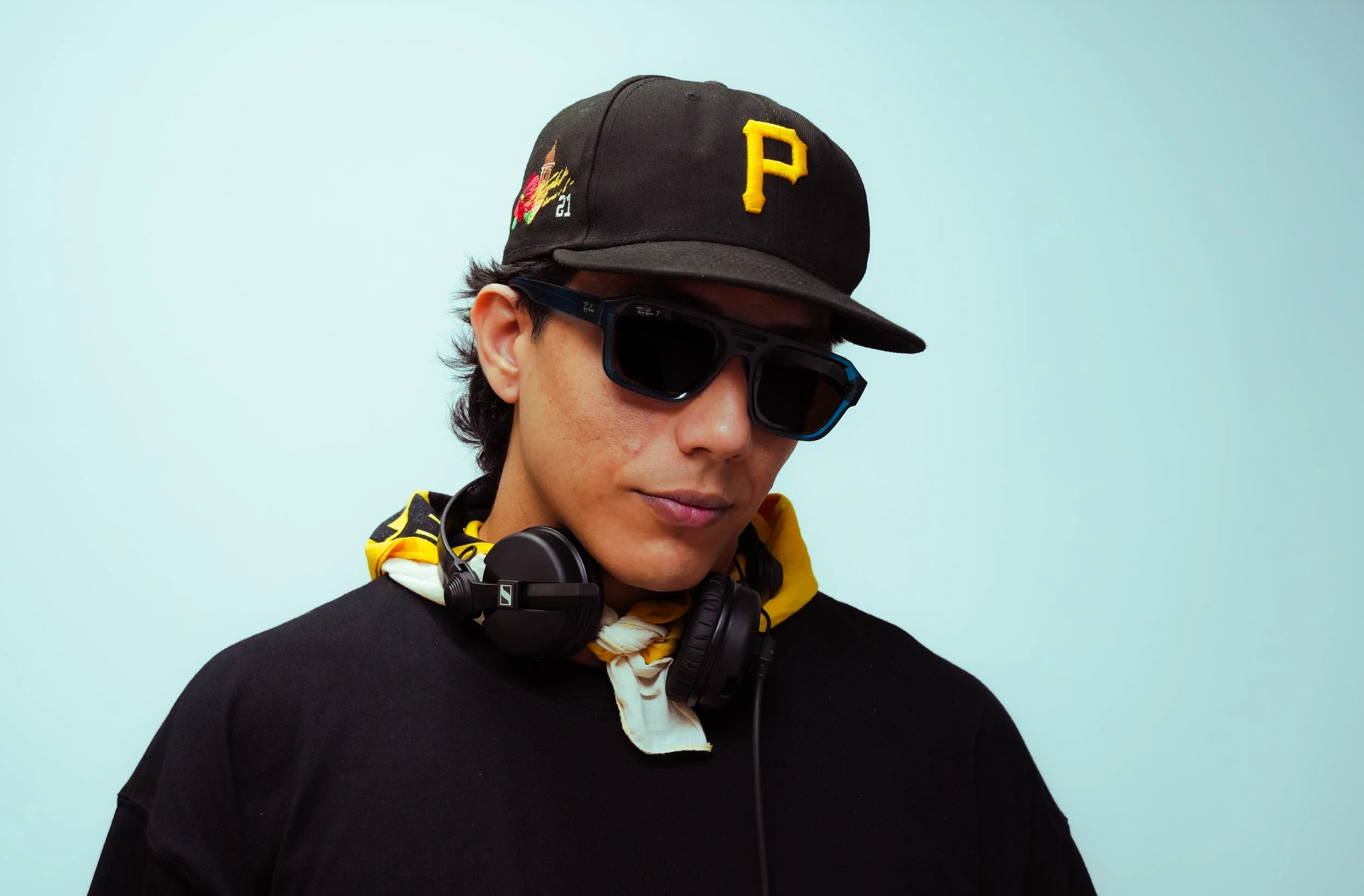 Young man wearing a black baseball cap with a yellow P, black sunglasses, and a black shirt, with headphones around his neck and a yellow bandana around his neck, against a light blue background.