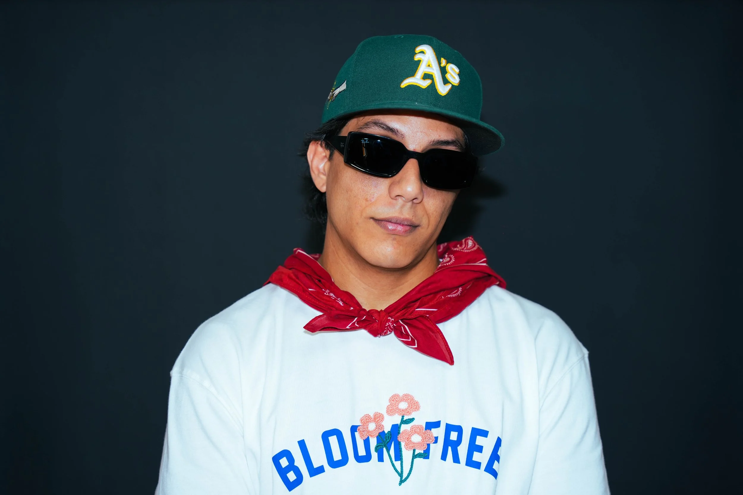 A young man wearing a green Oakland Athletics baseball cap, black sunglasses, a red bandana around his neck, and a white T-shirt with a graphic of pink flowers and the word 'BLOOMFREE' against a dark background.